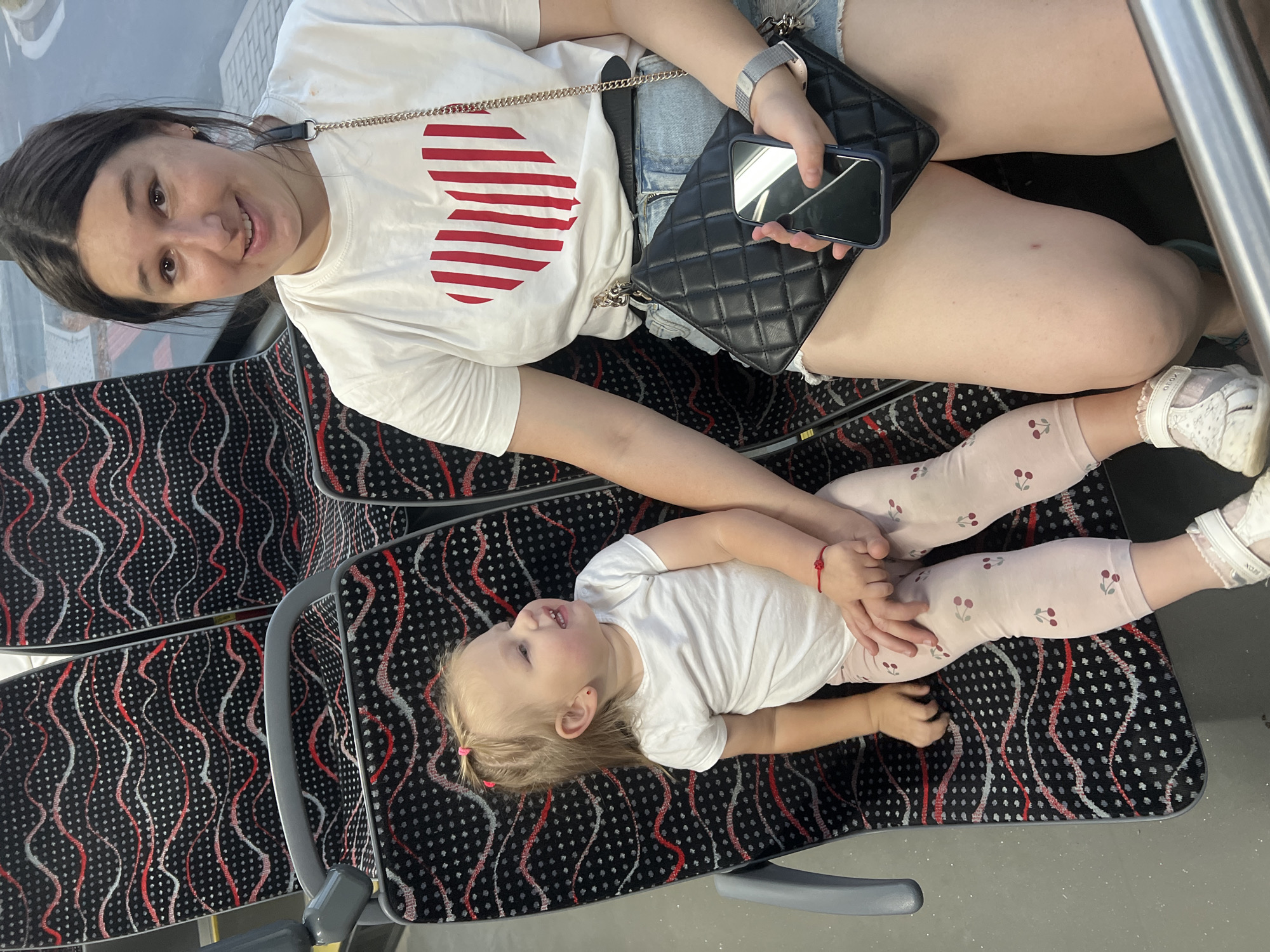 An adult female with dark hair is sitting on a bus seat next to a young child with blonde hair. The child is wearing a white t-shirt and light pink leggings with cherries. The adult female is wearing a white t-shirt with a red striped heart design and denim shorts. She is holding a black quilted purse and a smartphone. The bus seats have a black and red wavy pattern. The image appears to be taken on a public transportation vehicle, likely a bus or tram, in Pilsen, Czechia. The atmosphere is casual and everyday.