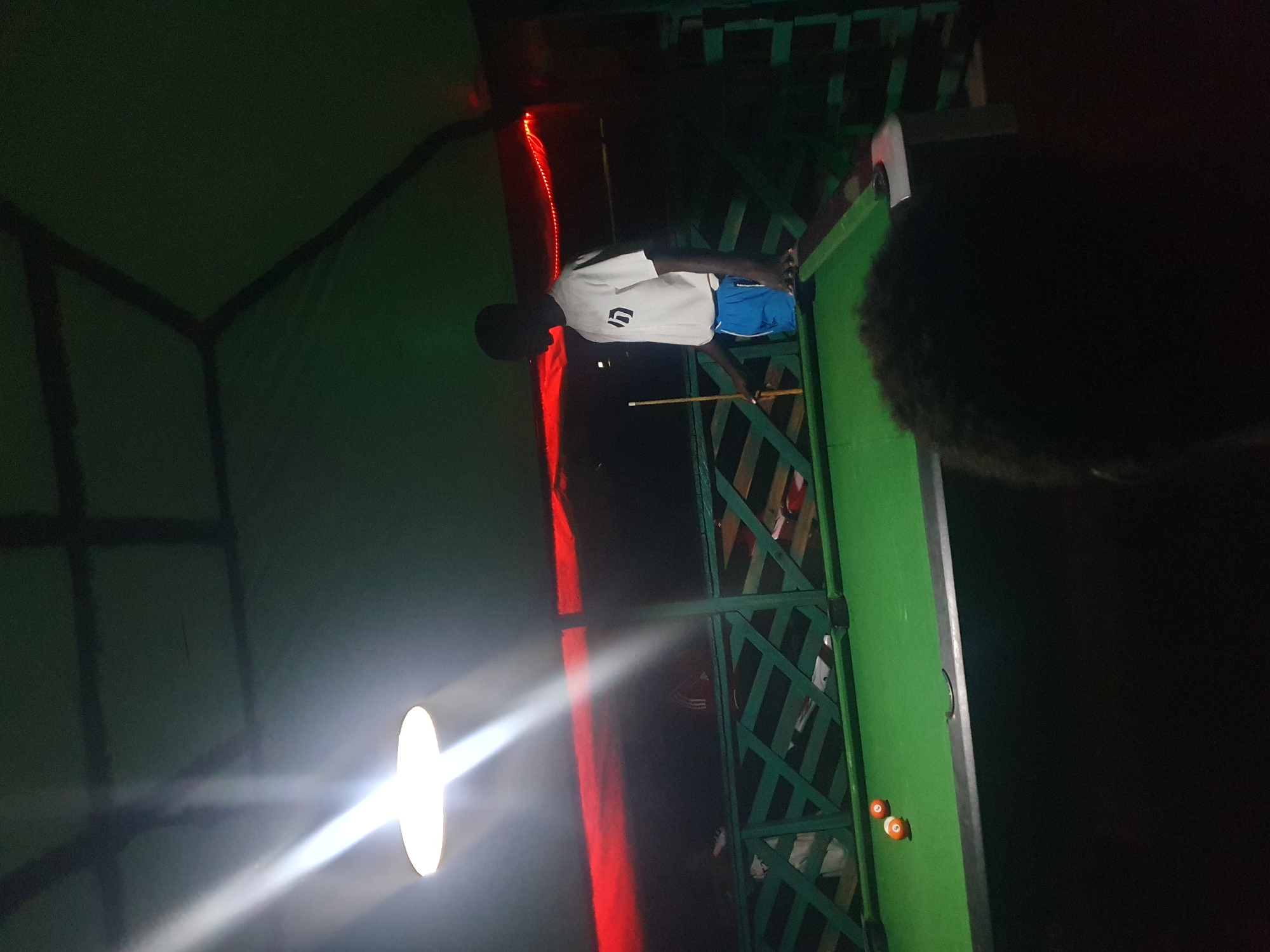In Lokoja, Nigeria, a dimly lit outdoor billiards area is depicted. A young man, silhouetted against a bright overhead light and red neon strip lighting, stands near a green pool table. He is wearing a white t-shirt with a graphic logo and blue shorts, holding a cue stick. Two orange billiard balls are positioned on the table. The background features a green wooden railing and a dark, possibly covered, ceiling. The scene appears to be a casual recreational space, possibly a bar or a private outdoor setup, with emphasis on the pool table and the solitary player. The lighting creates strong contrasts and shadows, contributing to a nocturnal atmosphere.