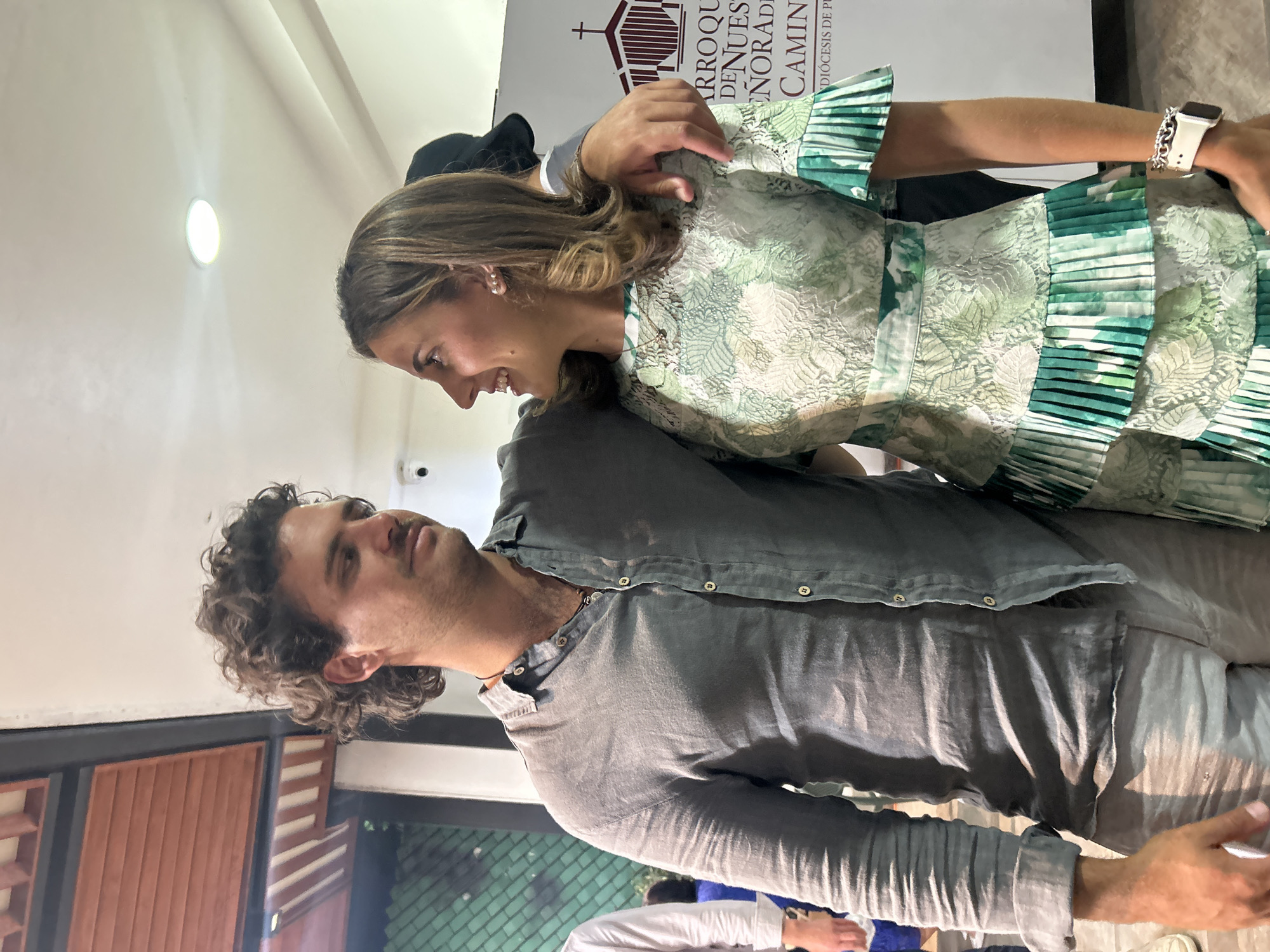 A woman in a green dress is interacting with a man in a gray shirt indoors. The background features a banner with religious symbols and text. The location is Unknown, Unknown.