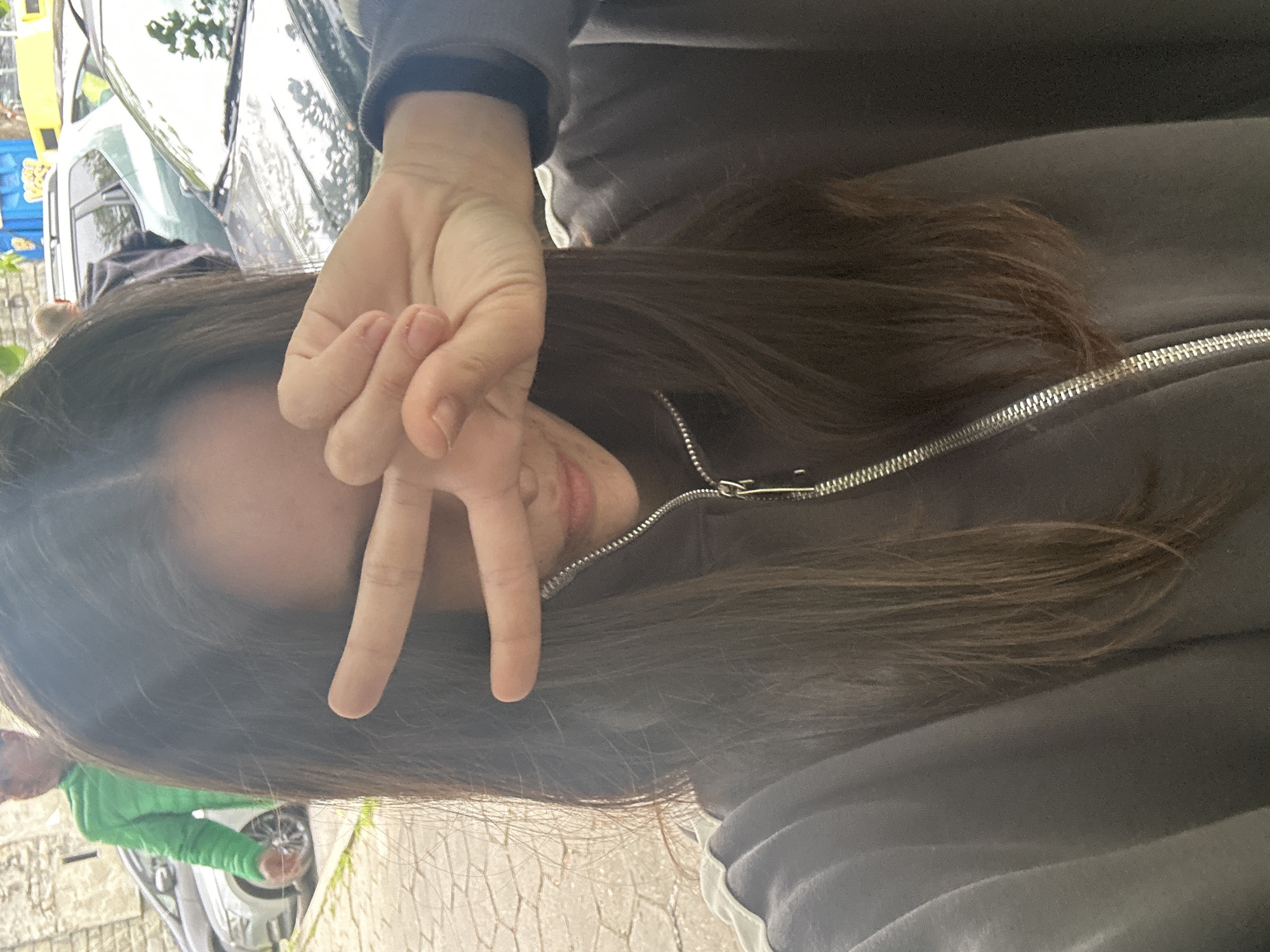 The image, taken in Veliko Tarnovo, Bulgaria, features a person (likely a young woman) in the foreground, capturing a selfie outdoors during daylight hours. The main subject's right hand is raised to her face, forming a V-sign with two fingers, which partially covers her left eye and cheek. She has long, straight, dark brown hair flowing over her shoulders and chest, and is wearing a dark-colored (possibly black or dark grey) zip-up hoodie with a prominent silver zipper. Her lips are subtly visible beneath her hand.
In the background, an outdoor street scene is visible. To the left, a person wearing a bright green jacket is partially seen, appearing to stand near a parked vehicle. Several cars are parked along what seems to be a paved street or pathway, with one light-colored car clearly visible. The ground is covered with light-toned, possibly cobblestone or patterned, paving. Reflections of trees and buildings are visible on the car windows, indicating an urban setting. The V-sign suggests a casual or friendly mood. There is no visible text in the image.