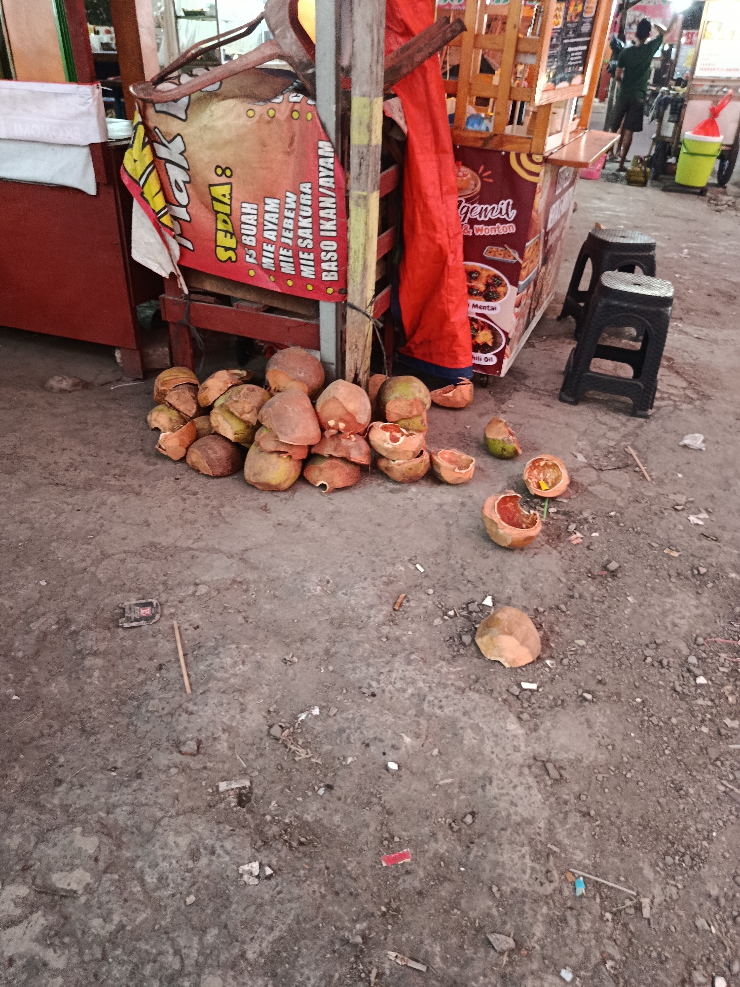 A small food cart is set up on a dusty, outdoor ground. A banner hangs from the cart, advertising various food items like mie ayam and mie jebew. In front of the cart, a pile of halved coconuts sits on the ground, some appearing to be fresh and others discarded. Two black plastic stools are placed near the cart. In the background, a person can be seen in silhouette, seemingly operating the food cart. The ground is littered with debris, including small pieces of paper and plastic.