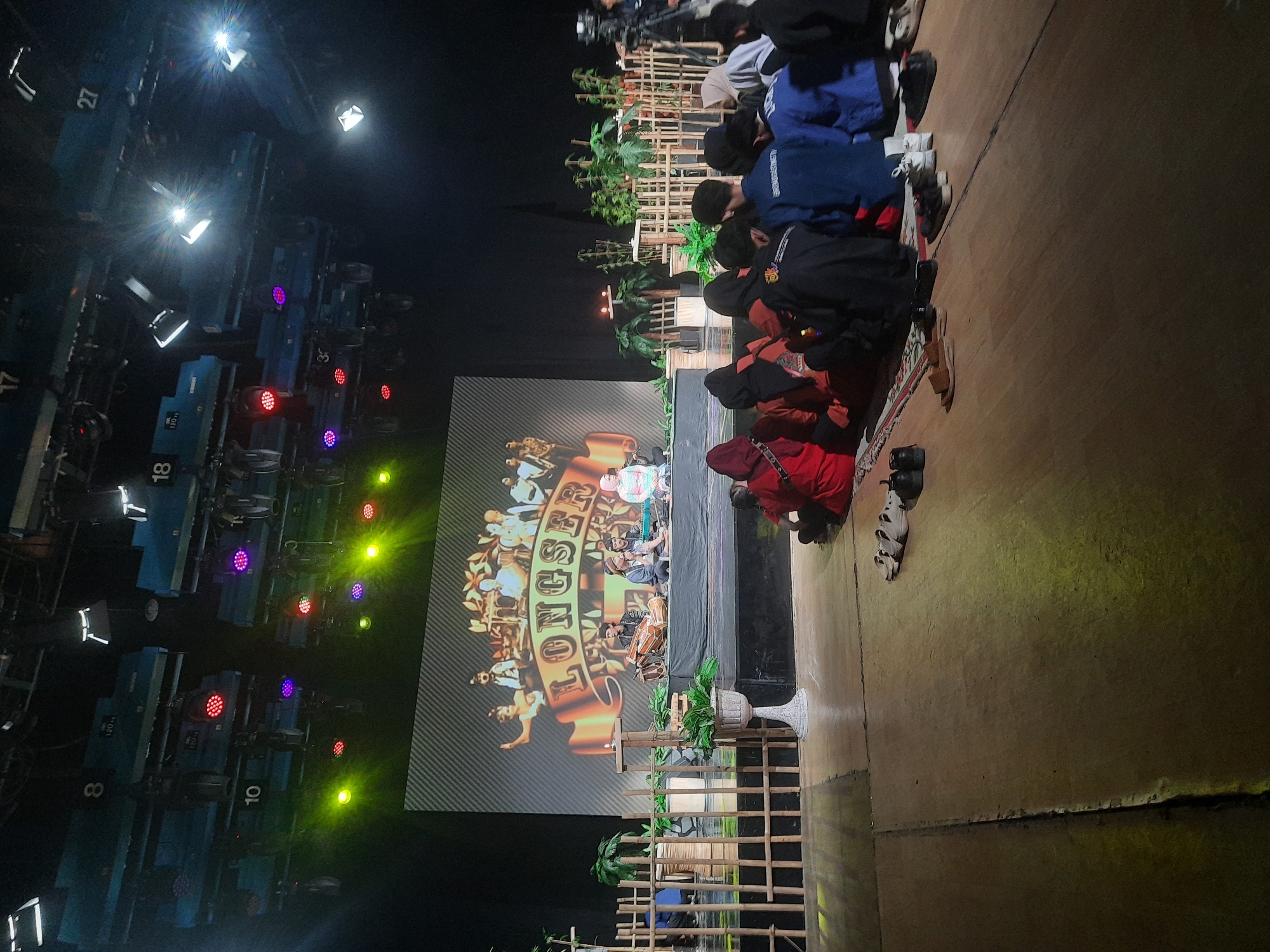 This is a medium shot, full-frame, eye-level photograph taken indoors, likely in a studio setting with stage lighting. The scene depicts a performance or presentation taking place on a raised stage, with a large screen displaying graphics behind the performers. In the foreground, a group of people are seated on the floor, facing the stage, with their backs to the camera.
The stage area is set up with traditional wooden fencing and potted plants, creating a rustic ambiance. On the stage, there are several individuals, possibly musicians or performers, seated and playing instruments. The screen behind them features an ornate logo that appears to read "LONGSER". The lighting is dramatic, with spotlights and colored lights illuminating the stage and casting yellow and green hues on the floor.
The group in the foreground consists of at least ten individuals, dressed in casual attire, some wearing headscarves. They are seated on a rug, and several pairs of shoes are placed on the floor around them. The environment suggests an indoor performance space, possibly a television studio or a cultural event venue. There are no identifiable landmarks that would place this scene specifically in Bandung City, Indonesia, beyond the user's request.
