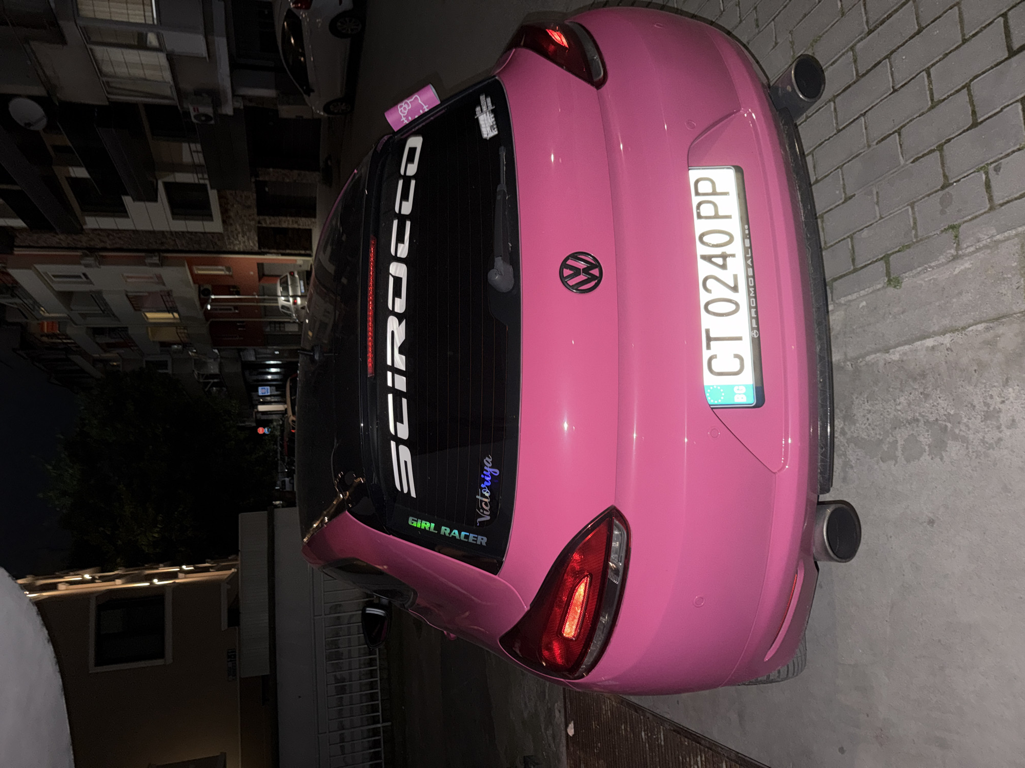 The image shows the rear of a bright pink Volkswagen Scirocco parked on a paved street at night in Plovdiv, Bulgaria. The car has a black roof and tinted rear windows. A sticker on the rear window reads "GIRL RACER" vertically, and a stylized text "Victoriya" below it. Another white sticker with a Hello Kitty design is visible on the right side of the rear window. The license plate of the car is clearly visible and reads "CT 0240 PP". The Volkswagen logo is present on the trunk lid. The background shows multi-story apartment buildings with some windows lit, and trees. The street is illuminated by streetlights.
