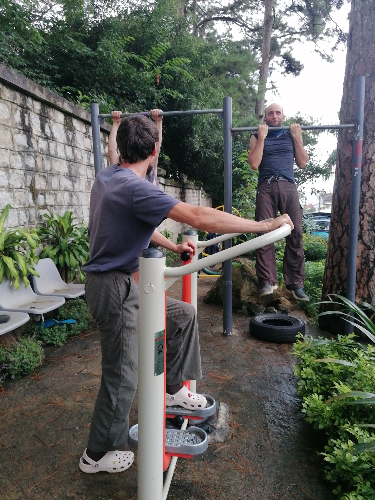 Two adult males are at an outdoor exercise area in Da Lat, Vietnam. One man, with short hair, is performing a pull-up on a horizontal bar. He is wearing a dark blue t-shirt and brown trousers. The second man, with dark hair, is standing on an exercise machine, facing away from the camera. He is wearing a grey t-shirt and grey trousers. He is holding onto a bar of the machine. The ground is wet, suggesting it has rained recently. There are trees and green foliage surrounding the exercise area, with a stone wall in the background.
