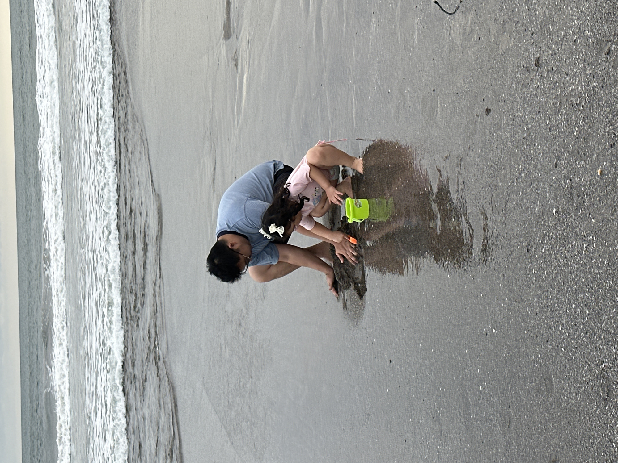 In North Kuta, Indonesia, a father and daughter are playing on the beach. The father, wearing a blue t-shirt and glasses, is crouched down, while his daughter, in a pink t-shirt and hair bow, is squatting beside him. They are both interacting with the wet sand, with the daughter holding a small orange shovel and a bright green bucket. The ocean waves are gently rolling onto the shore behind them, creating a serene and natural backdrop. The sand is dark and wet, with visible reflections of the figures in the shallow water.
