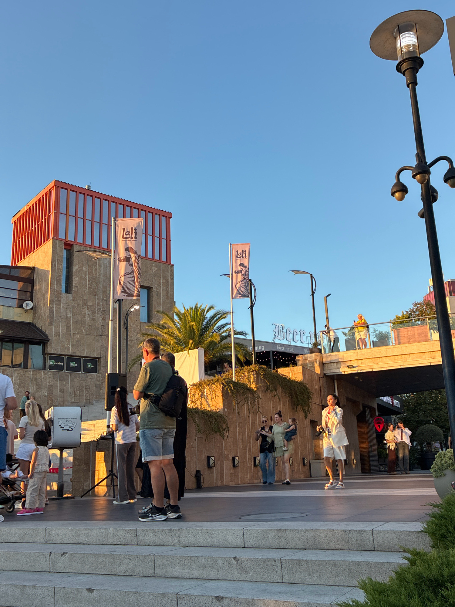 An outdoor scene at dusk on what appears to be a promenade or public square in Sochi, Russia, features a variety of people gathered. The architecture includes a modern building with a red-trimmed upper section and stone-clad lower levels. Two vertical banners with stylized figures and Cyrillic text hang from flagpoles in front of the building. A palm tree is visible behind the banners.
Several groups of people are scattered across the scene. In the foreground, a man in a green t-shirt and shorts stands with a backpack, facing away from the camera. Beside him, a woman in a striped t-shirt and dark pants also stands. Further to the left, a child in a light-colored outfit stands near a stroller.
On a slightly elevated walkway or bridge in the background, a woman in a white jacket and shorts is holding a microphone, suggesting she might be performing or addressing a crowd. Other individuals are visible on this bridge and in the background near buildings with signage. The sky is a clear blue, transitioning to a warmer hue near the horizon, indicating sunset. Streetlights are present, one a tall, modern design with multiple lamps. Lush greenery, including ivy-like plants and potted bushes, adds to the environment.