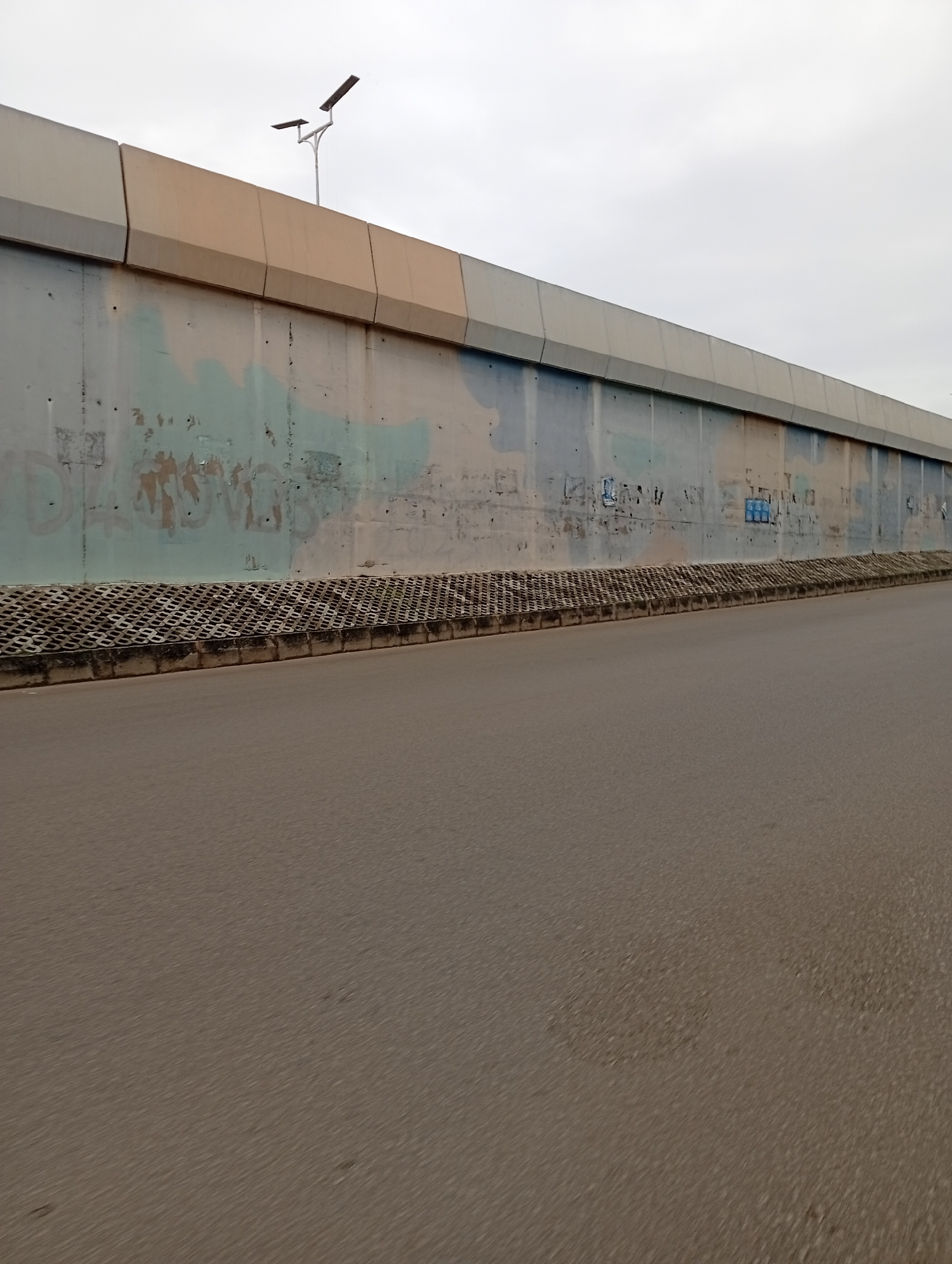 The image shows a roadside view of a long, concrete retaining wall, likely part of an overpass or elevated road. The wall has a beige-colored cap along the top edge, and below this, the concrete surface is worn and shows signs of graffiti, with faded blue and brown markings. A band of interlocking concrete blocks runs along the base of the wall, serving as a drainage or retaining structure. In the background, the sky is overcast, suggesting a cloudy or possibly rainy day. A single solar-powered street light with two panels is visible mounted on a pole near the top of the wall. The foreground is dominated by a paved road, which appears to be asphalt and shows signs of wear and tear, with some darker patches and embedded debris. The perspective suggests the photo was taken from a moving vehicle. There is no discernible human activity or event taking place in the scene. The overall impression is of an urban or semi-urban infrastructure setting under a dull sky. No clear text is legible, although some graffiti is present on the wall. The location context is Jos, Nigeria.