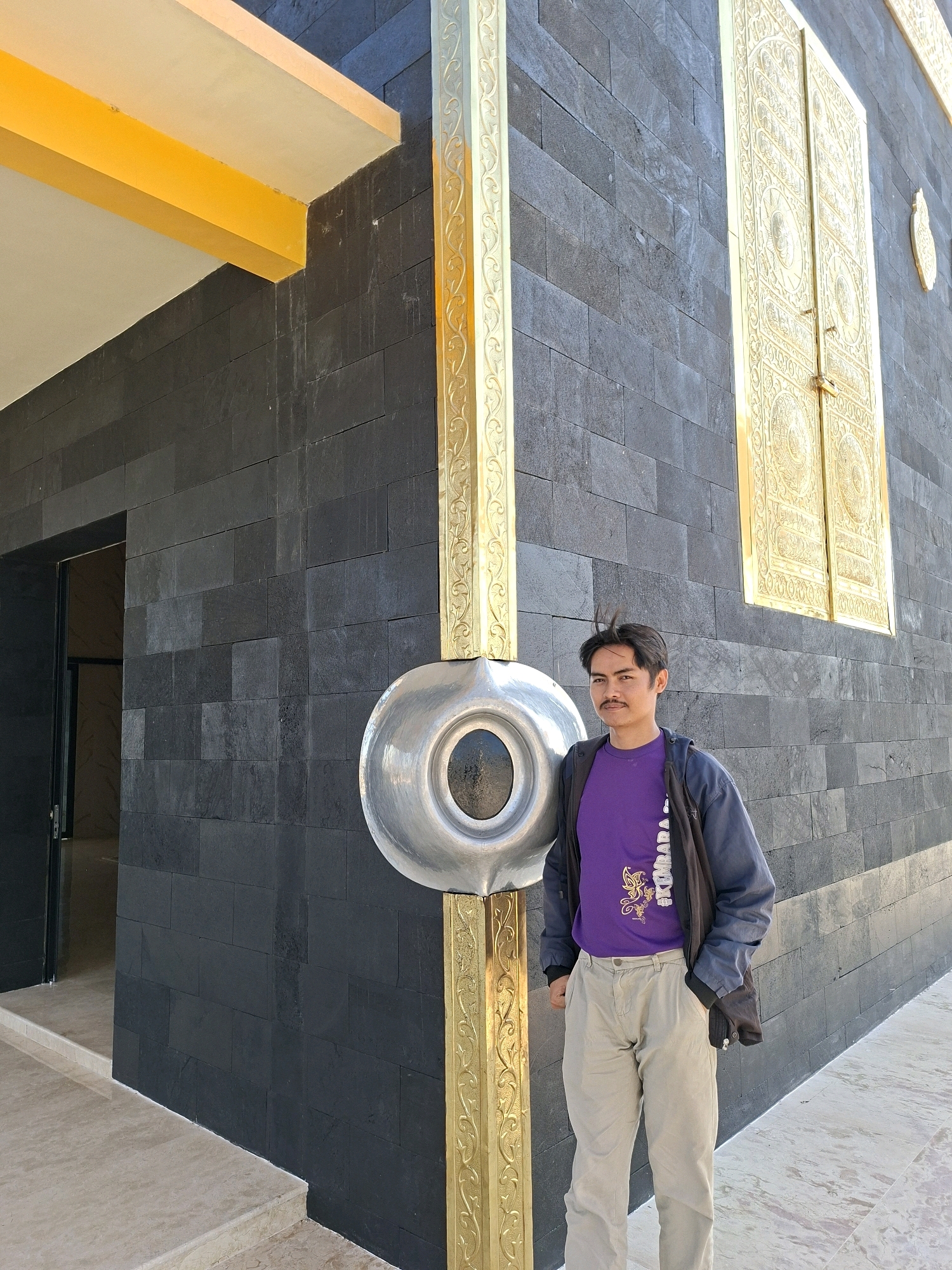 A medium shot captures a young man standing outside a dark stone building with gold embellishments in Jrengik, Indonesia. The man, with dark hair styled forward and a mustache, wears a purple t-shirt with a gold graphic, tan pants, and a dark jacket draped over his shoulders. He stands to the right of a circular silver object mounted on a gold column. The building features large, ornate gold double doors to the right of the man. A yellow overhang is visible on the left side of the building. The ground is paved with light-colored stone.