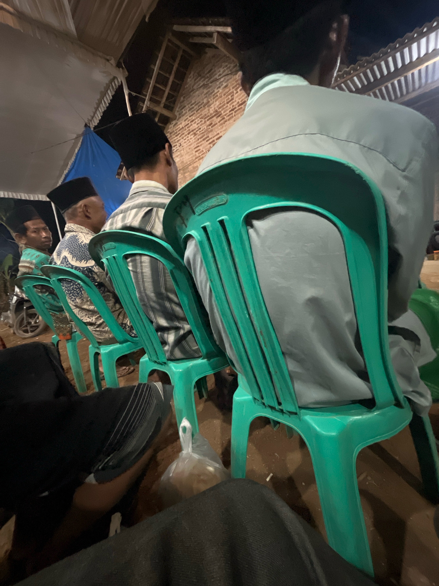 The image depicts an outdoor gathering at night, possibly in Kebumen, Indonesia. The scene is dominated by several individuals seated on bright green plastic chairs, facing away from the camera. They appear to be men, most wearing black peci hats, a traditional cap worn by Muslim men in Indonesia. Their attire includes patterned shirts and plain grey clothing.
In the background, a temporary structure with a white tarp and a blue tarp is visible, suggesting an event or gathering. A brick wall and wooden beams are also part of the background, indicating a rustic or semi-constructed environment. A motorcycle is partially visible on the left side of the frame, and a clear plastic bag containing some items rests on the ground. The overall lighting suggests it is nighttime, with artificial lights illuminating the area. The perspective is from a seated position, looking at the backs of the attendees.