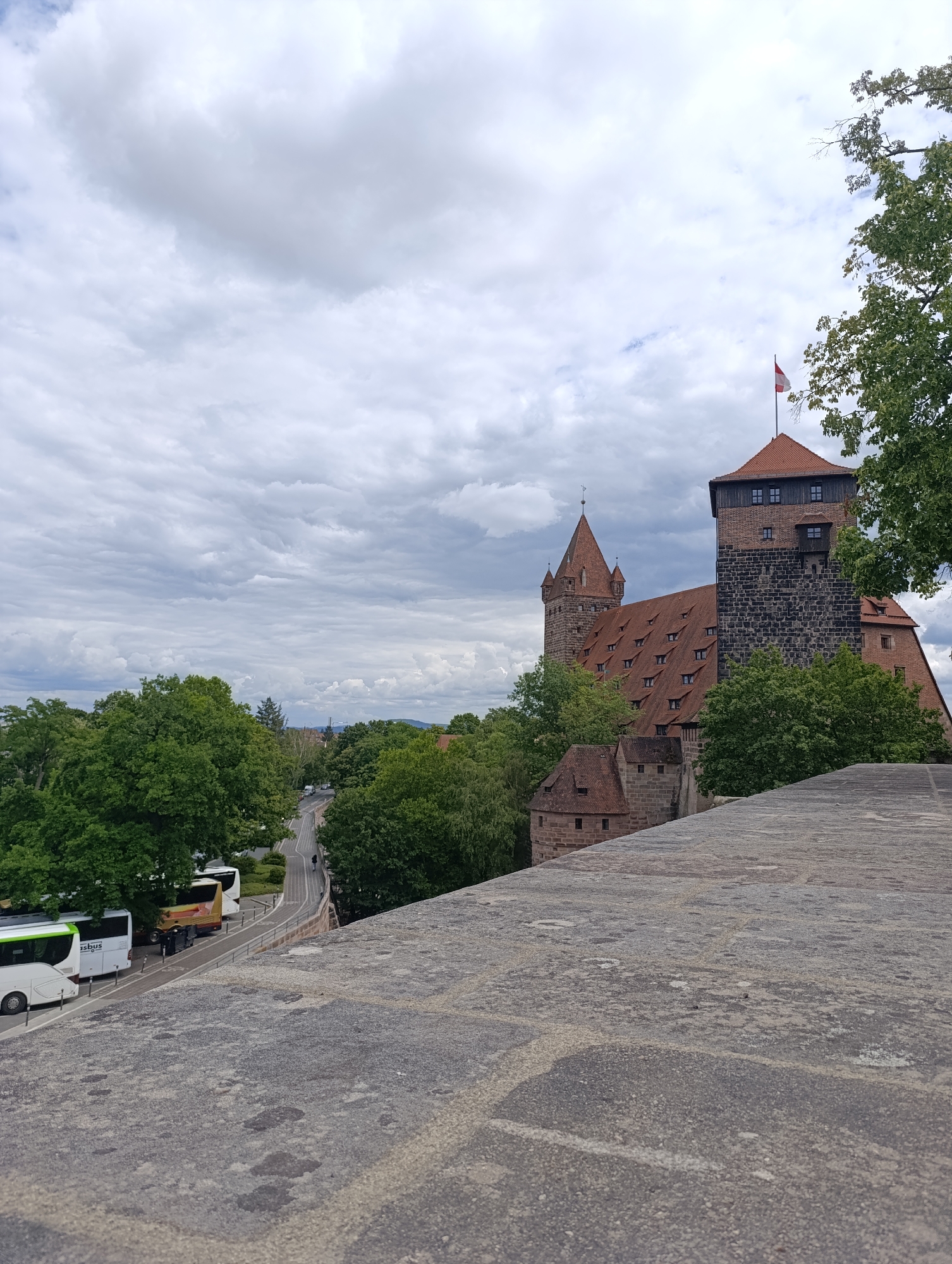 This is a medium shot taken from a raised vantage point overlooking the Imperial Castle of Nuremberg. The castle, a prominent historical landmark in Nuremberg, Germany, is visible on the right side of the image. It features multiple towers with red tiled roofs and dark stone construction. A flag, red and white, flies from the highest tower.
In the middle ground, a road curves around a hill, lined with lush green trees. Several white and yellow buses are parked along the side of the road, suggesting a tourist area or transportation hub. A few figures can be seen walking on the road.
The foreground is dominated by a textured stone surface, possibly a wall or walkway, leading the eye towards the castle. The sky is heavily clouded with a mix of white and gray formations, indicating an overcast day. The overall scene depicts a section of the historic city, blending architectural heritage with the natural landscape and signs of modern activity.