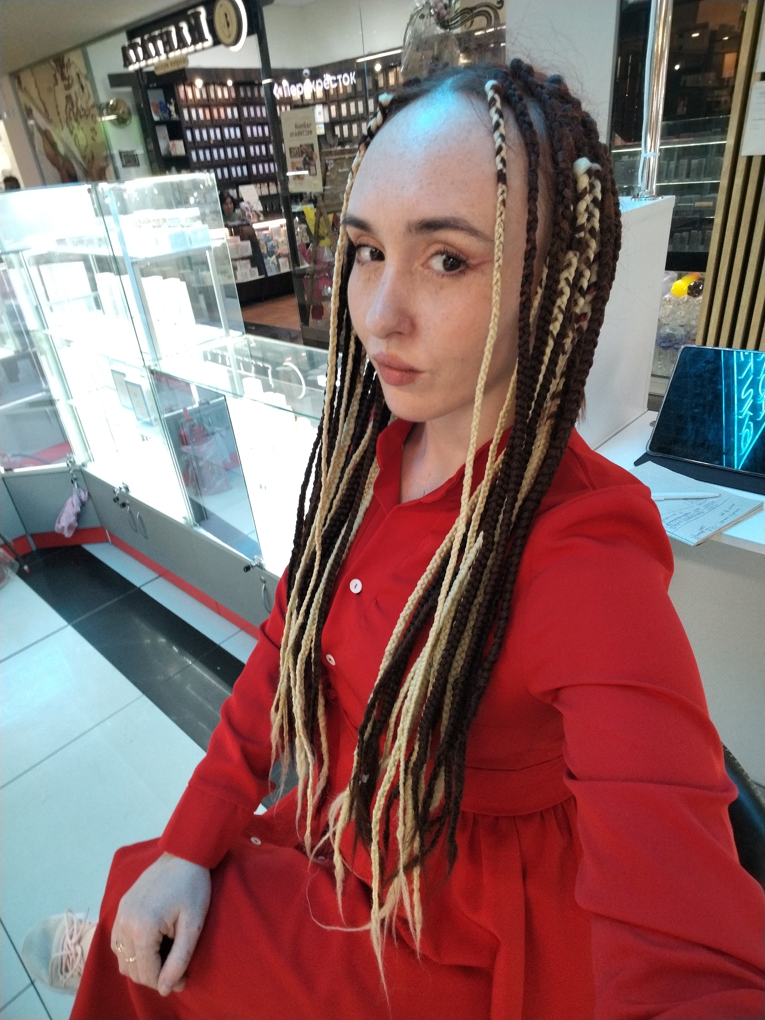 A woman with light skin, freckles, and braided hair in brown and blonde strands sits in what appears to be a shopping mall or commercial space. She is wearing a long-sleeved red dress. The background includes a display case, a store with shelves filled with merchandise, and general mall architecture. A computer tablet and some papers are visible on a white surface next to her. The store in the background has signage in Russian including the word "ПЕРЕКРЁСТОК" which suggests the location is in Russia, potentially in the city of Izhevsk, based on the store's prevalence in that region.
