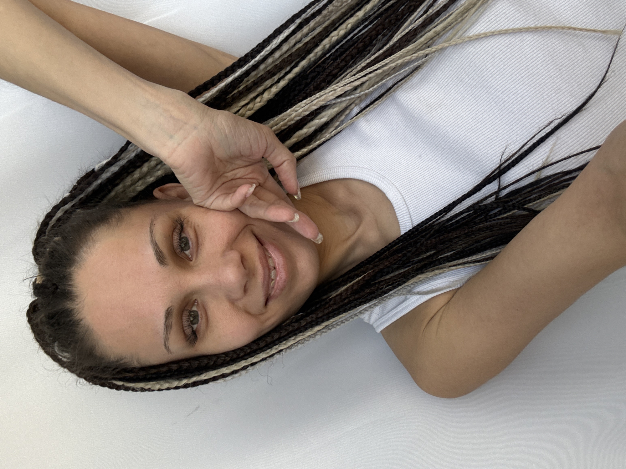 A woman with brown eyes and long, braided hair, some of which is blonde, lies on a white surface. She is wearing a white top and has her hand near her face, smiling. The location is not provided.