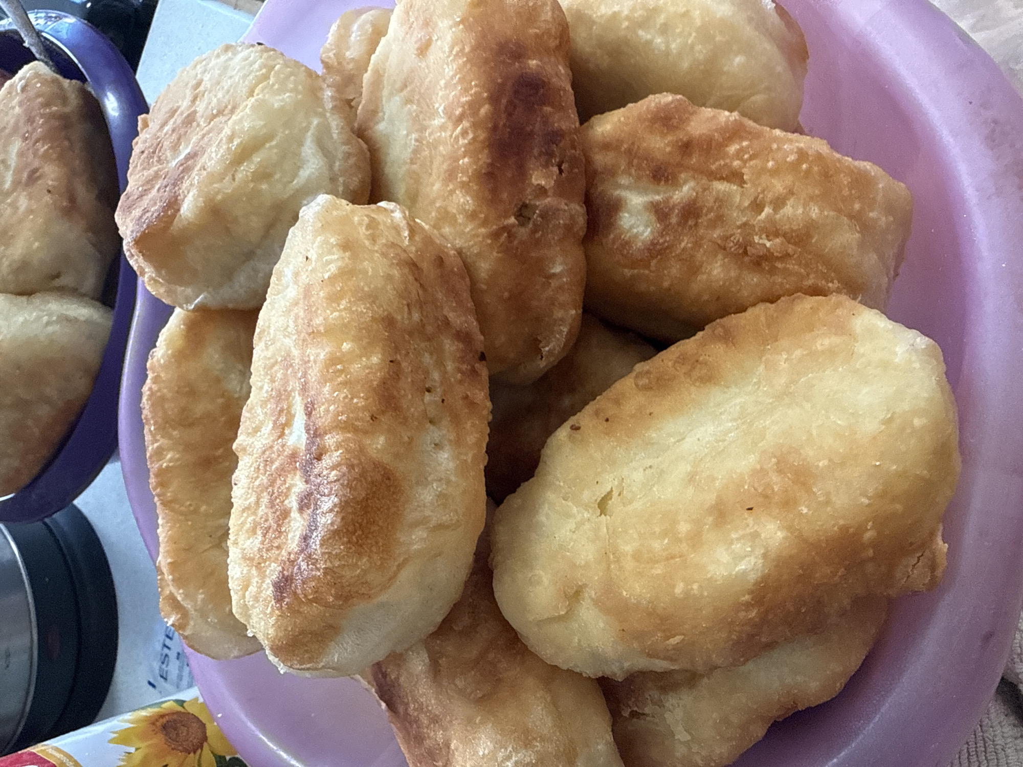 A close-up shot shows a pile of golden-brown fried pastries in a purple bowl. The pastries appear soft and pillowy. A second bowl with similar pastries sits to the left of the purple bowl. A box with a sunflower design sits in the lower left corner of the frame. The location and people in this image are indeterminable, as the context does not provide clues.