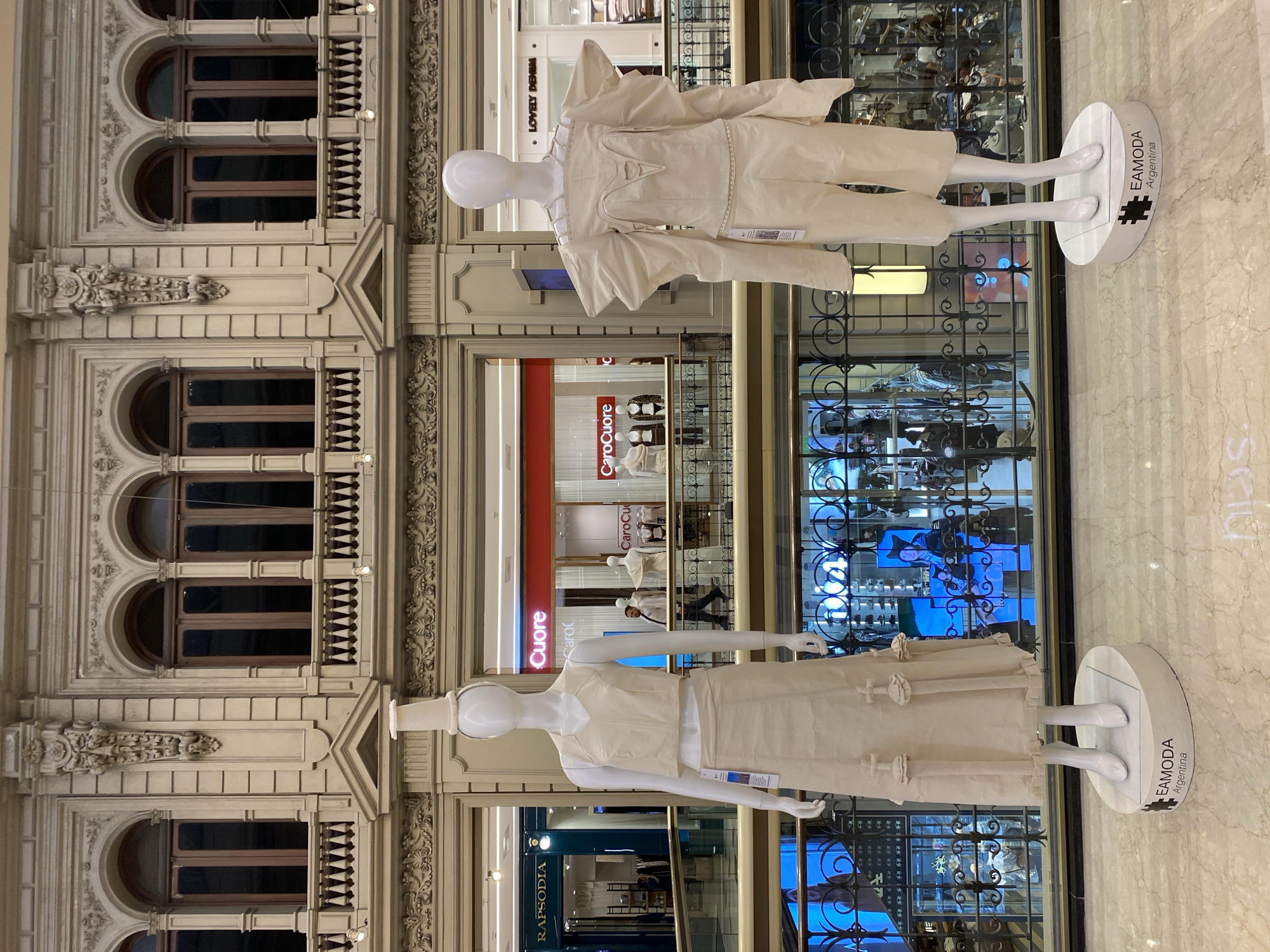 The image captures an interior scene at Galerías Pacífico in Buenos Aires, Argentina. Two mannequins stand on pedestals labeled "EAMODA Argentina," displaying minimalist off-white outfits. They are positioned near an ornate iron railing overlooking other stores in the shopping center. In the background, "Caro Cuore" and "Rapsodia" storefronts are visible on the lower levels. The architectural elements of Galerías Pacífico, including arched windows and decorative molding, add to the scene's visual complexity. The floor is made of marble.