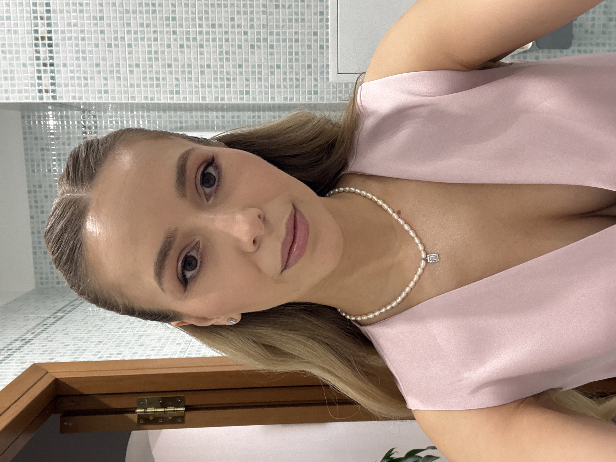 A woman with blonde hair is taking a selfie in a bathroom in Moscow, Russia. She is wearing a light pink dress and a pearl necklace with a small square pendant. Her makeup is natural, with a focus on her eyes and lips. The background shows a tiled wall and a wooden cabinet. The lighting suggests it could be daytime or artificial lighting is being used. The woman's expression is neutral, and the overall mood is relaxed. There is no visible text in the image.