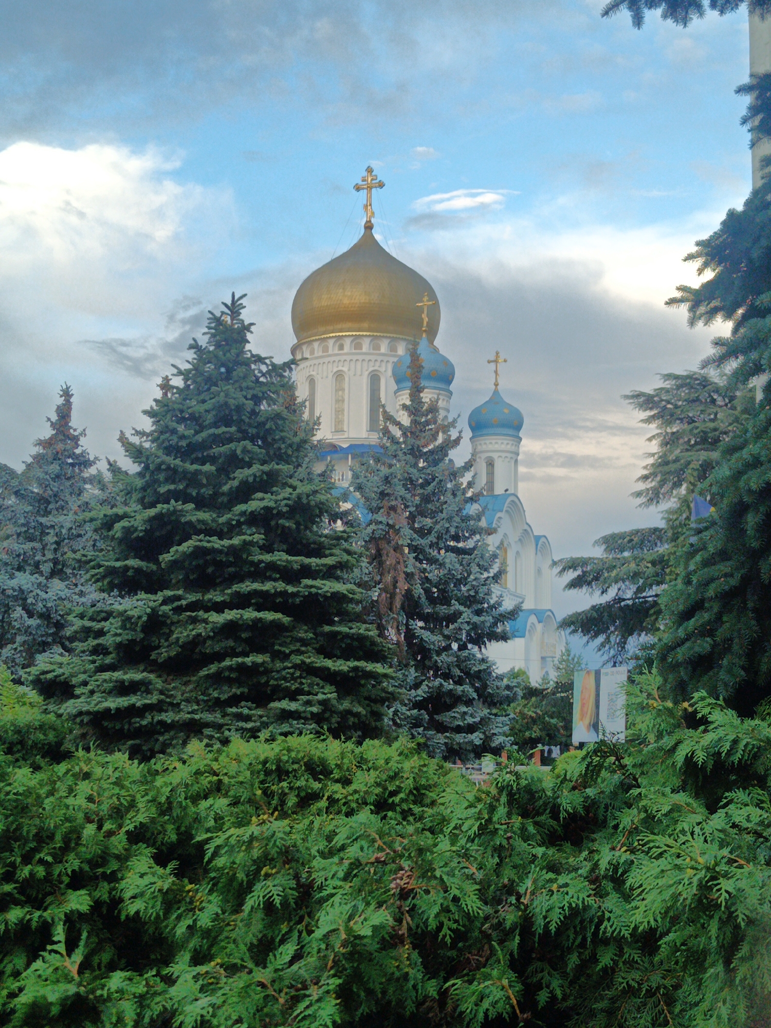 The image shows the Church of the Transfiguration in Uzhhorod, Ukraine, on a cloudy day. The church features a large golden dome topped with a cross, and several smaller blue domes also topped with crosses. The white building of the church has arched windows. In the foreground, there are numerous dense evergreen trees, obscuring the lower part of the church and its immediate surroundings. The sky is overcast with scattered clouds.