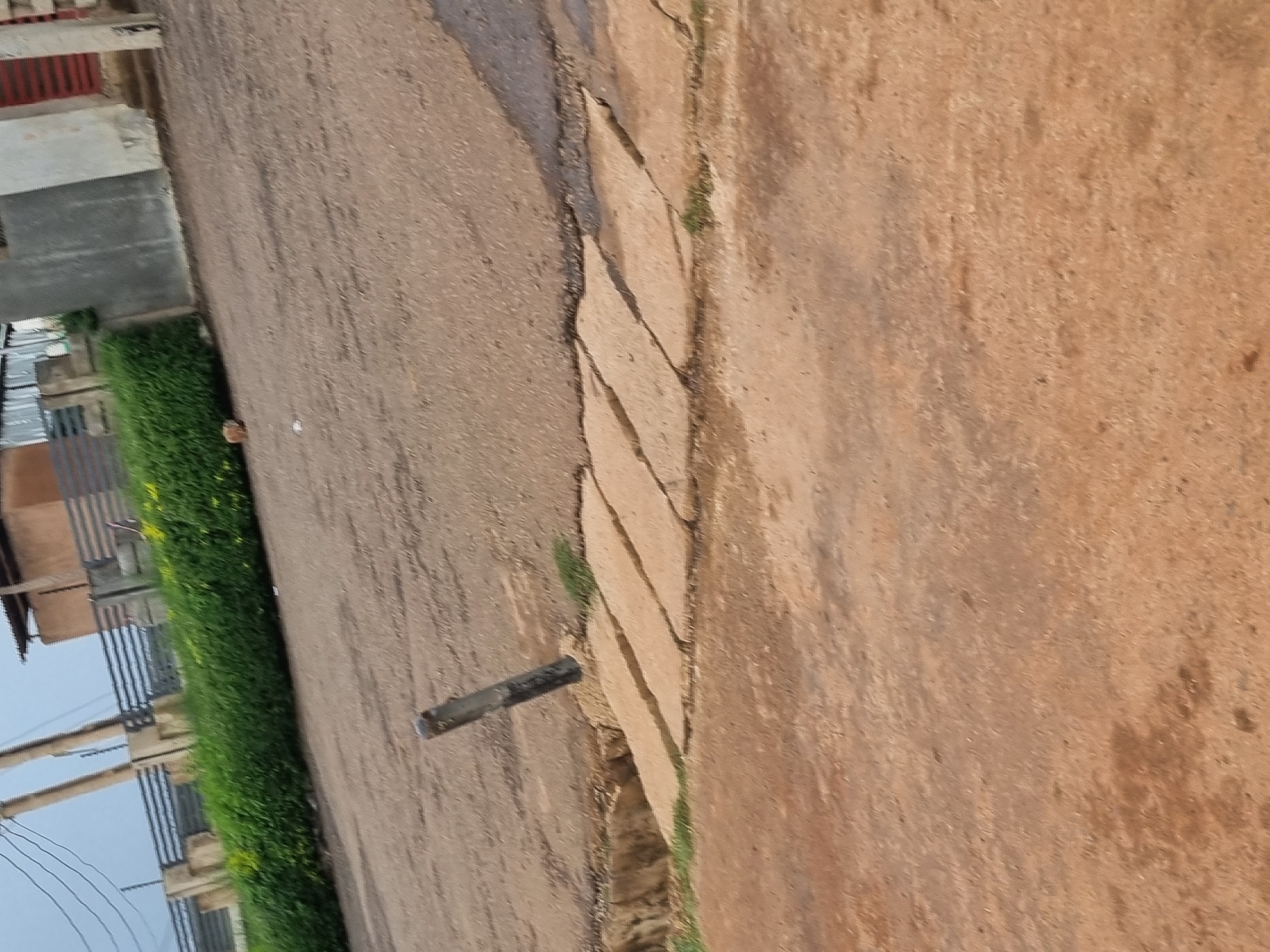 The image depicts an outdoor scene, likely a street or roadside area in Jos, Nigeria. In the foreground, a dirt path with patches of dry grass is visible. A gray, broken concrete structure, possibly part of a drainage system or a broken pathway, runs diagonally across the mid-ground. A dark, upright object, possibly a metal post or pipe, juts out from the broken concrete. Beyond this, a paved road with visible cracks and uneven sections extends towards the background. The background features a dense green hedge, beyond which a fence and some buildings with corrugated metal roofing and concrete structures are discernible. Utility poles with overhead wires are also visible in the upper left portion of the image, suggesting an urban or semi-urban environment. The overall scene appears weathered and somewhat neglected, consistent with an outdoor environment in a city like Jos.