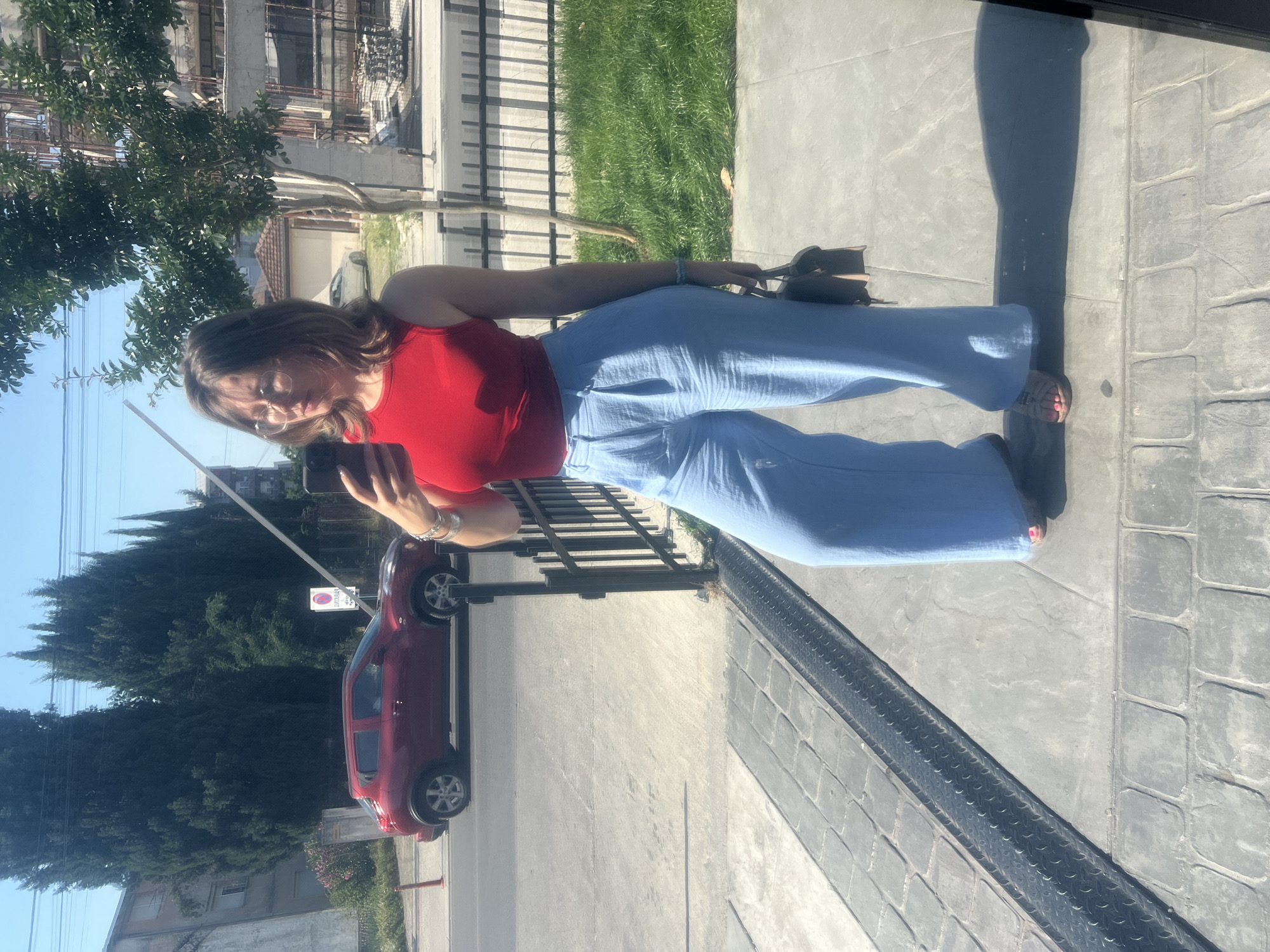 A woman stands outdoors on a paved surface next to a low metal fence, holding a smartphone and appearing to take a selfie. She wears a red top and light blue pants. Behind her is a street with a red car parked on the side. Trees and buildings are visible in the background. The location is Podgorica, Montenegro.