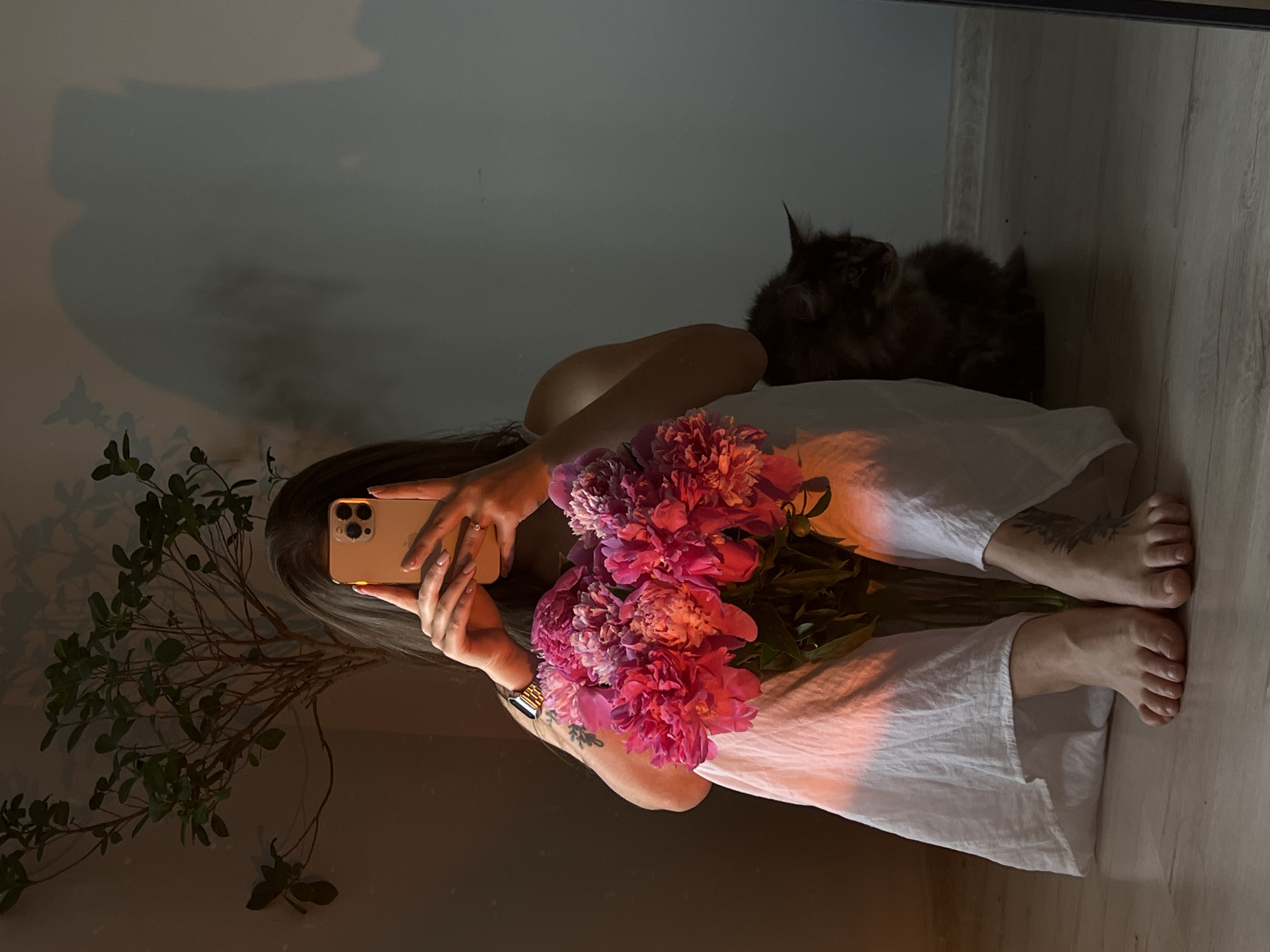 A person is sitting on the floor in a room, holding a bouquet of pink peonies and a smartphone. The person is wearing a white dress. A cat is sitting next to the person, looking to the right. There is a plant in the background. The lighting suggests it is either early morning or late afternoon. The image was taken indoors, likely in a residential setting. The location is not specified in the image, and therefore cannot be identified as being in Ternopil, Ukraine.