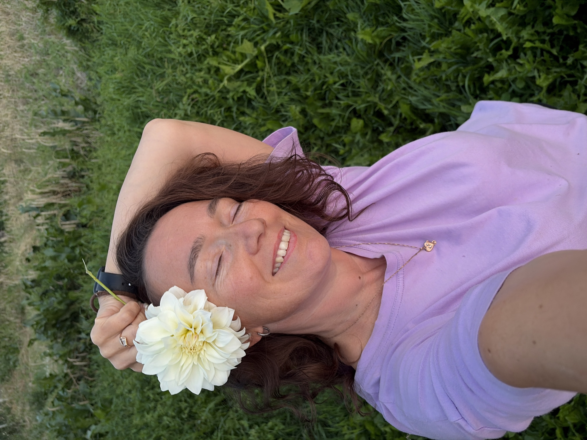 A woman with brown curly hair is captured in a selfie, holding a white dahlia above her head with her right hand. She is wearing a light purple t-shirt and a necklace with a small pendant. Her eyes are closed, and she has a relaxed smile. Her left arm is bent, resting behind her head. The background consists of lush green grass and foliage. The location is not specified as Olkhovka, Russia, as there is no visual evidence to support this claim.