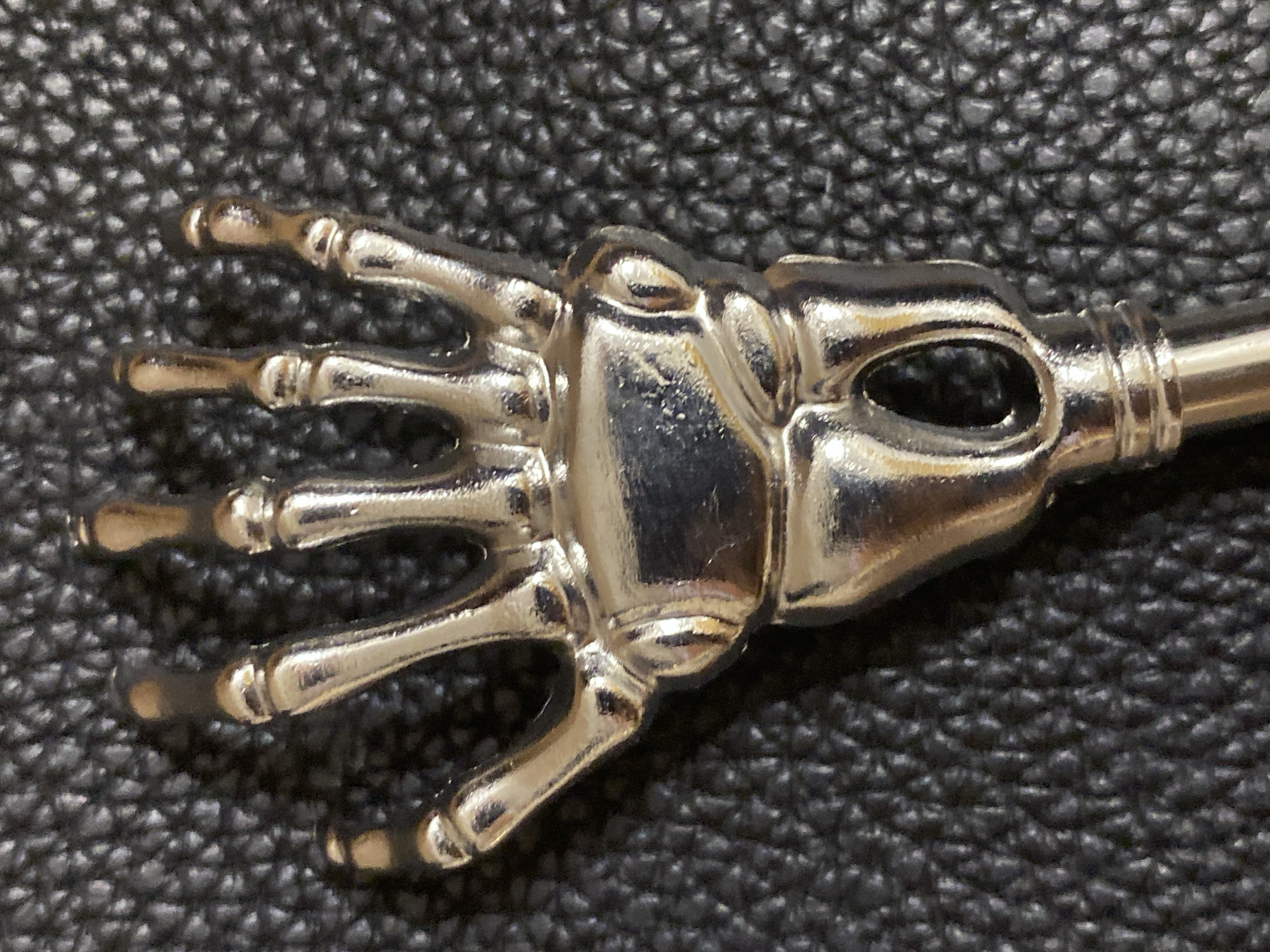 A close-up shot shows a silver-colored skeleton hand-shaped bottle opener resting on a dark, textured surface, possibly leather. The hand, with its distinct skeletal fingers and metacarpals, lies horizontally with the fingers pointing left. The surface upon which the opener rests is dark and grainy. No indication of location.