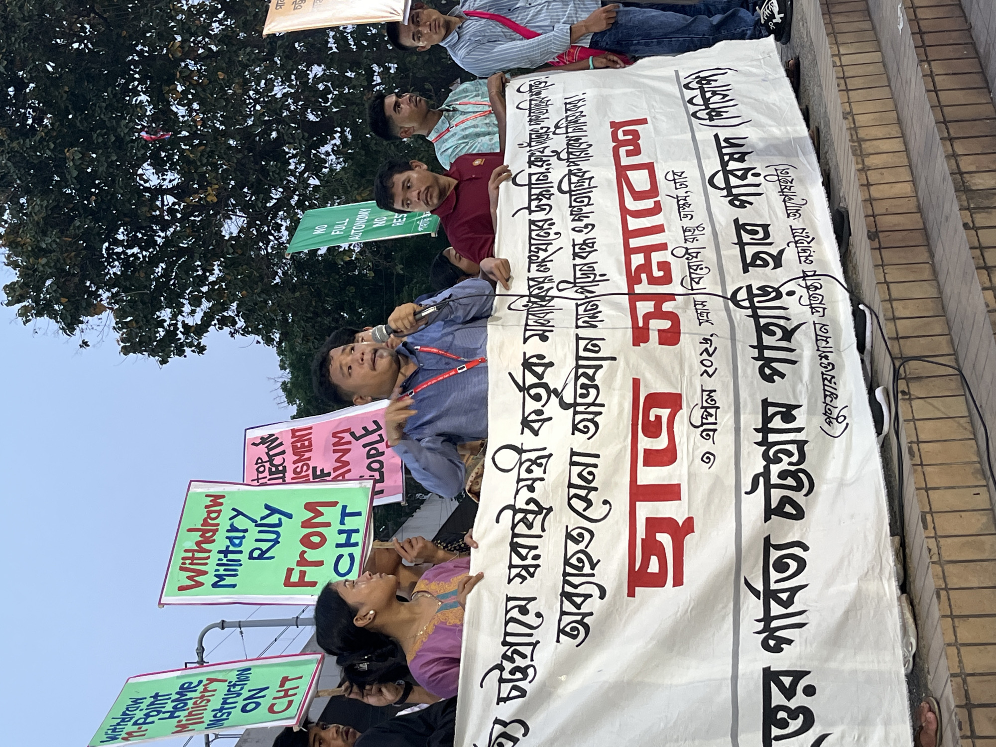 A street-level photograph captures a demonstration in Dhaka, Bangladesh. The event, identified as a "Student Rally," features individuals holding protest signs and a large banner. Some of the signs are written in English, demanding the withdrawal of military rule from CHT (Chittagong Hill Tracts) and an end to the Home Ministry's instruction on CHT. The primary banner, in Bengali, calls for an end to military campaigns, particularly after the Minister of Home Affairs. A man with a microphone addresses the crowd. The protest appears to be occurring in a public area with trees visible in the background, adjacent to stairs. The rally is dated April 3, 2026, near the anti-terrorism sculpture at the University of Dhaka (Dhaka Bishwabidyalaya). The title “Chatra Somabesh” translates to “Student Assembly.” The banner further names the organization as the "Parbatya Chattagram Pahari Chhatra Parishad (PCP)", indicating a student organization focused on the Chittagong Hill Tracts.