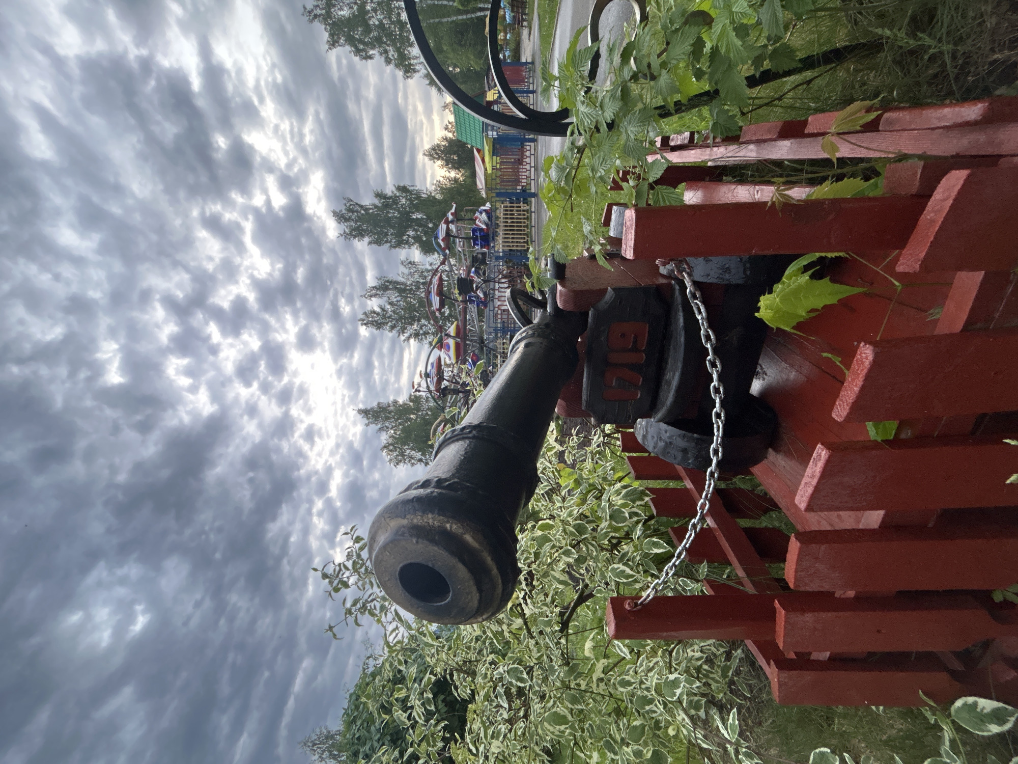 A ground-level, outdoor shot depicts a cannon exhibit in Chaikovsky, Russia. The cannon is positioned on a red wooden structure with a chain attached. Green foliage and trees surround the exhibit, and in the background are amusement park rides. The sky is cloudy.