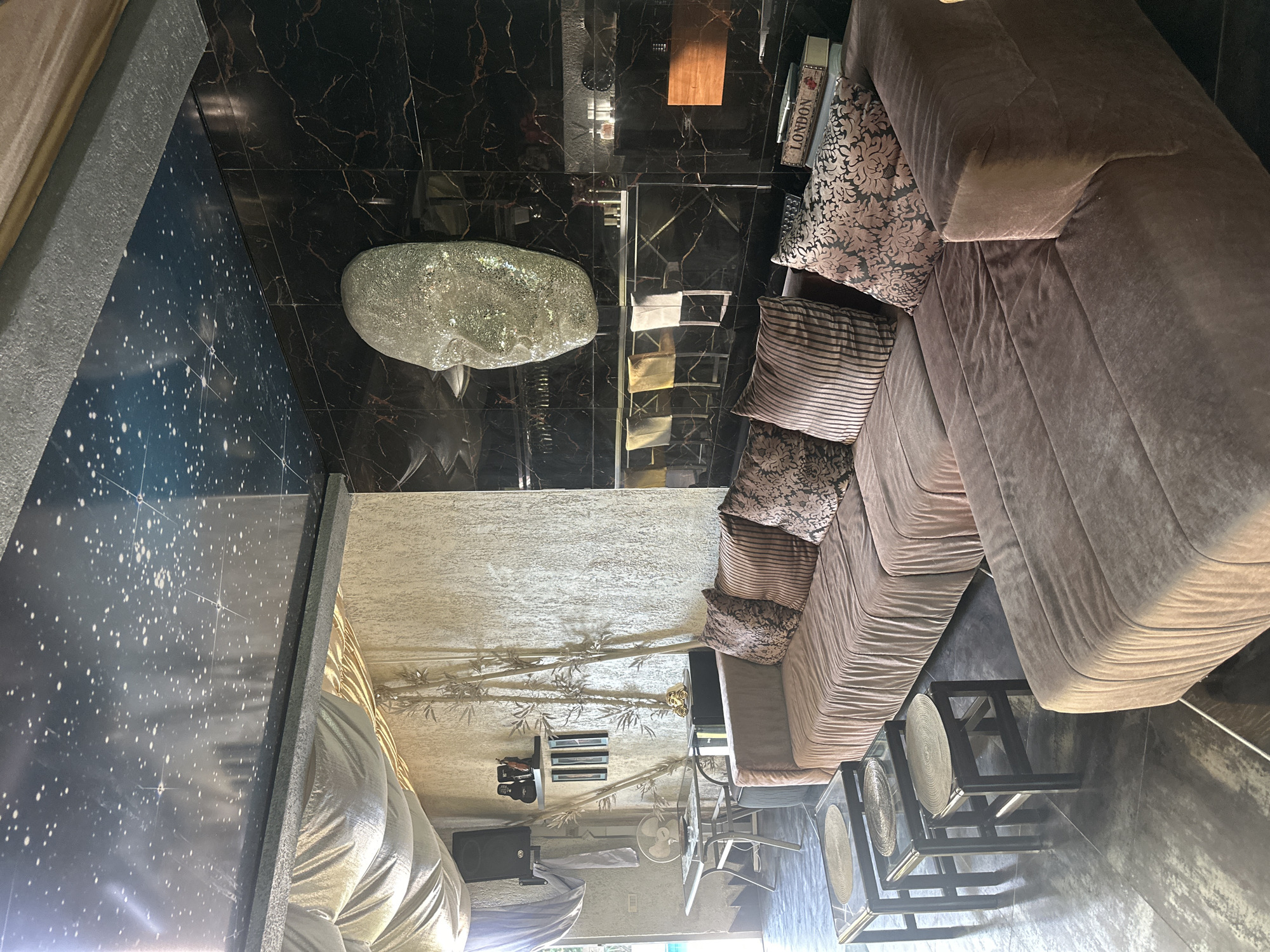 The image depicts a luxurious interior space, possibly a lounge or living room, in Brca, Montenegro. The room features a large, sectional sofa upholstered in a taupe velvet fabric, adorned with several decorative throw pillows in various patterns and textures. In front of the sofa, a set of three nested square glass-top tables with dark wood frames are arranged on a dark, reflective floor.
The backdrop of the room is striking. The ceiling is adorned with a dark, glossy finish patterned with white stars, resembling a night sky. One wall is covered in large, black marble tiles with prominent gold veining, creating a dramatic effect. Mounted on this wall is a large, textured sculpture of a human face, made of a reflective, possibly glitter-encrusted material. Adjacent to the marble wall is a textured, light-colored wall, decorated with several tall, stylized bamboo-like elements in a golden hue. On a shelf mounted on this wall are a few decorative items, including what appear to be speakers, a small golden figurine, and framed artwork. Further down this wall, a glass-top table with a metal frame is set up, with a fan and other objects on its surface, suggesting it might be a workspace or a functional area.
The lighting in the room is atmospheric, with a warm glow emanating from a wooden-mounted light fixture on the marble wall, and other ambient lighting sources not directly visible. The overall impression is one of opulence and modern, eclectic design.