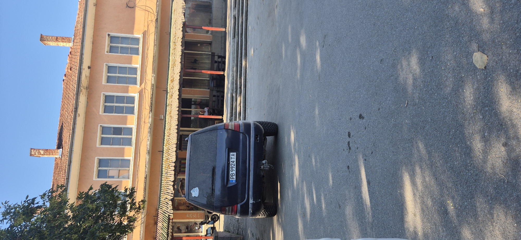 A dark blue Mercedes-Benz SUV is parked in an asphalt lot in front of a two-story building with an orange facade. The SUV's rear license plate reads "PB 9924 TA". The building has a tiled roof and multiple windows on both floors. A covered outdoor seating area with red-painted support beams is attached to the front of the building, with tables and benches visible. A person is sitting at one of the tables. A scooter is parked to the left of the building. Trees and sunlight cast shadows on the ground. The sky is clear and blue.