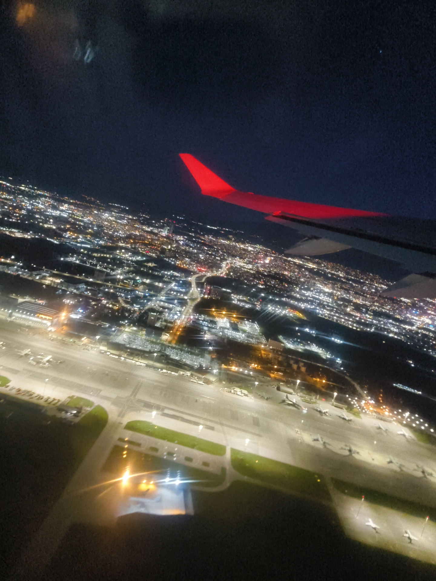 View from an airplane window at night. A red wing is visible in the upper right of the frame. Below, a sprawling cityscape is visible with numerous lights. In the foreground, an airport tarmac is seen with parked airplanes and illuminated runways.