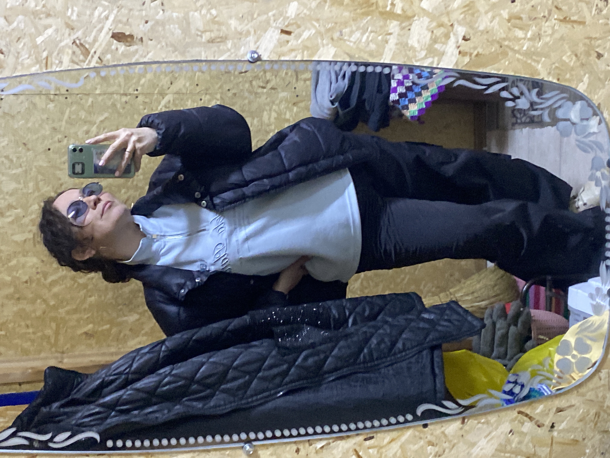 A person is taking a mirror selfie indoors. The background is a wall made of wooden panels. Clothes are hanging behind the mirror. The person is wearing sunglasses, a black puffer jacket, and a light-colored hoodie. They are holding a smartphone in their right hand. A black quilted jacket is in the lower left corner. The location is possibly Ol'khovskoye, Russia.