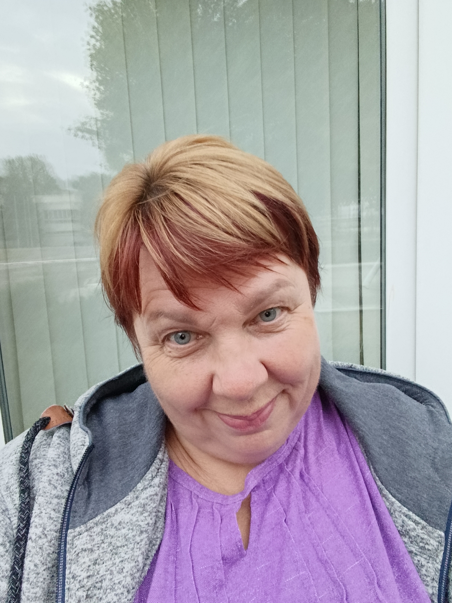 A medium-shot, eye-level selfie is taken outdoors, likely in the city of Svietlahorsk, Belarus, judging by the context of the image. A woman with short, layered blonde hair with red undertones is centered in the frame, facing the camera with a slight smile and a playful expression. She has bright blue eyes and fair skin with visible wrinkles around her eyes and mouth. She is wearing a purple V-neck top with a textured fabric, layered under a grey zippered hoodie. The background consists of vertical blinds, possibly on a window or glass door, with a muted reflection of trees and an overcast sky. The lighting is soft and diffused, suggesting an outdoor setting on a cloudy day.