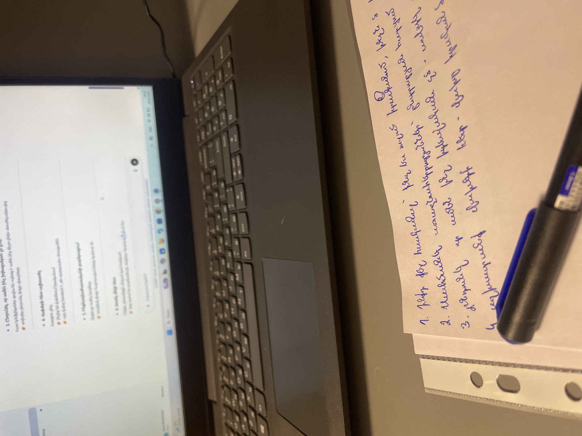 The image depicts a desk with a laptop, a notebook, and a pen. The laptop screen displays Armenian text, suggesting the user may be studying or working in Armenian. The notebook contains handwritten notes in Armenian. A black pen rests on top of the notebook. The scene is indoors. There is no specific information available to indicate the location as Yerevan, Armenia, or any specific details about the environment, people or interactions.