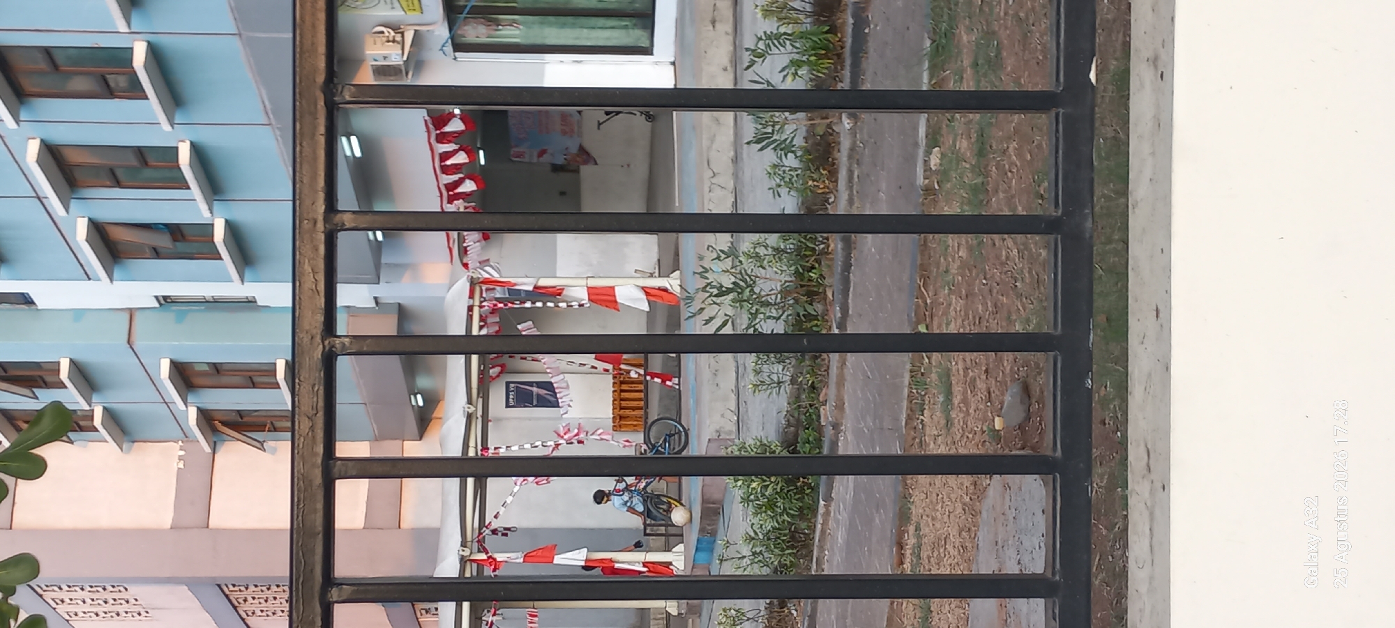 The image shows a young boy sitting on a blue bicycle next to a white ball, with a black metal fence in the foreground. The boy is wearing a light blue shirt and dark pants. Behind him, there is a building with light blue and white walls and several windows with white frames. Red and white triangular flags are strung across the front of the building. The ground is paved with concrete and dirt, with some sparse green plants. In the lower right corner of the image, text reads "Galaxy A32 25 Agustus 2026 17.28". The context of East Jakarta, Indonesia, is provided by the text overlay suggesting the date and possibly the location of capture.