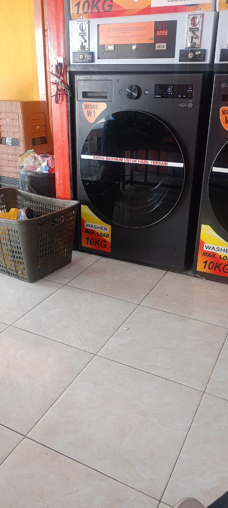 The image shows a row of commercial LG washing machines in what appears to be a self-service laundry establishment in Mekar Rahayu, Indonesia. The washing machines are predominantly black with large glass doors. Each machine has a label indicating "WASHER MAX. LOAD 10KG". Above the machines, a sign reads "10KG" in large red lettering on an orange background. To the left of the washing machines, there are stacked brown plastic chairs and a dark grey laundry basket filled with plastic bags containing various items, possibly detergent or other laundry supplies. A pair of scissors is attached to a red-painted pole or door frame to the left of the washing machines. The floor is tiled with light-colored square tiles. The overall environment suggests a functional, public space designed for laundry services.