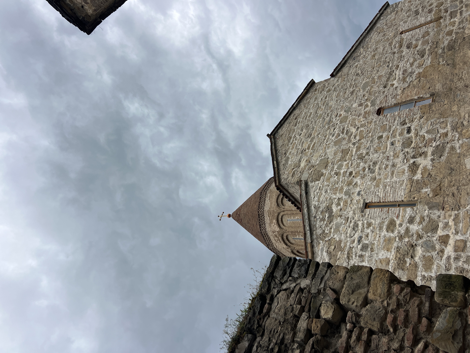 A low-angle shot captures the dome of a stone church in Shalikiantkari, Georgia, against a cloudy, overcast sky. The dome is conical with a tiled roof and topped with a cross. The church walls are constructed of rough-hewn stones, with narrow windows providing glimpses of the interior. To the left of the church, a crumbling stone wall, possibly part of an older structure, is visible. The overall impression is of an ancient, weathered building under a dramatic sky.