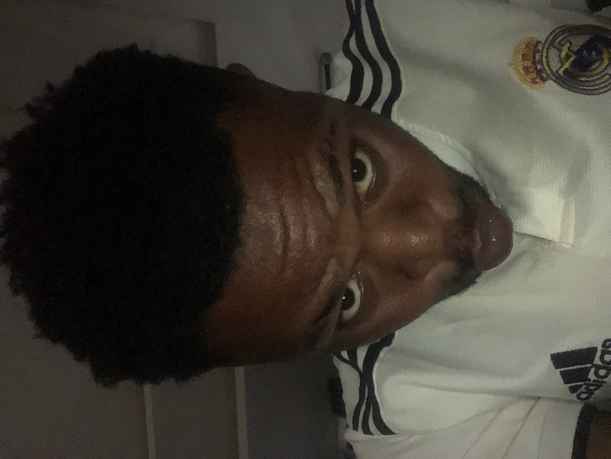 A close-up of a person's face and upper torso is shown in a rotated image. The individual has dark skin, dark curly hair, and appears to be wearing a white athletic jersey with black stripes and a Real Madrid emblem. The person's face appears slightly sweaty. The background is indistinct. Location: Unknown, Unknown.