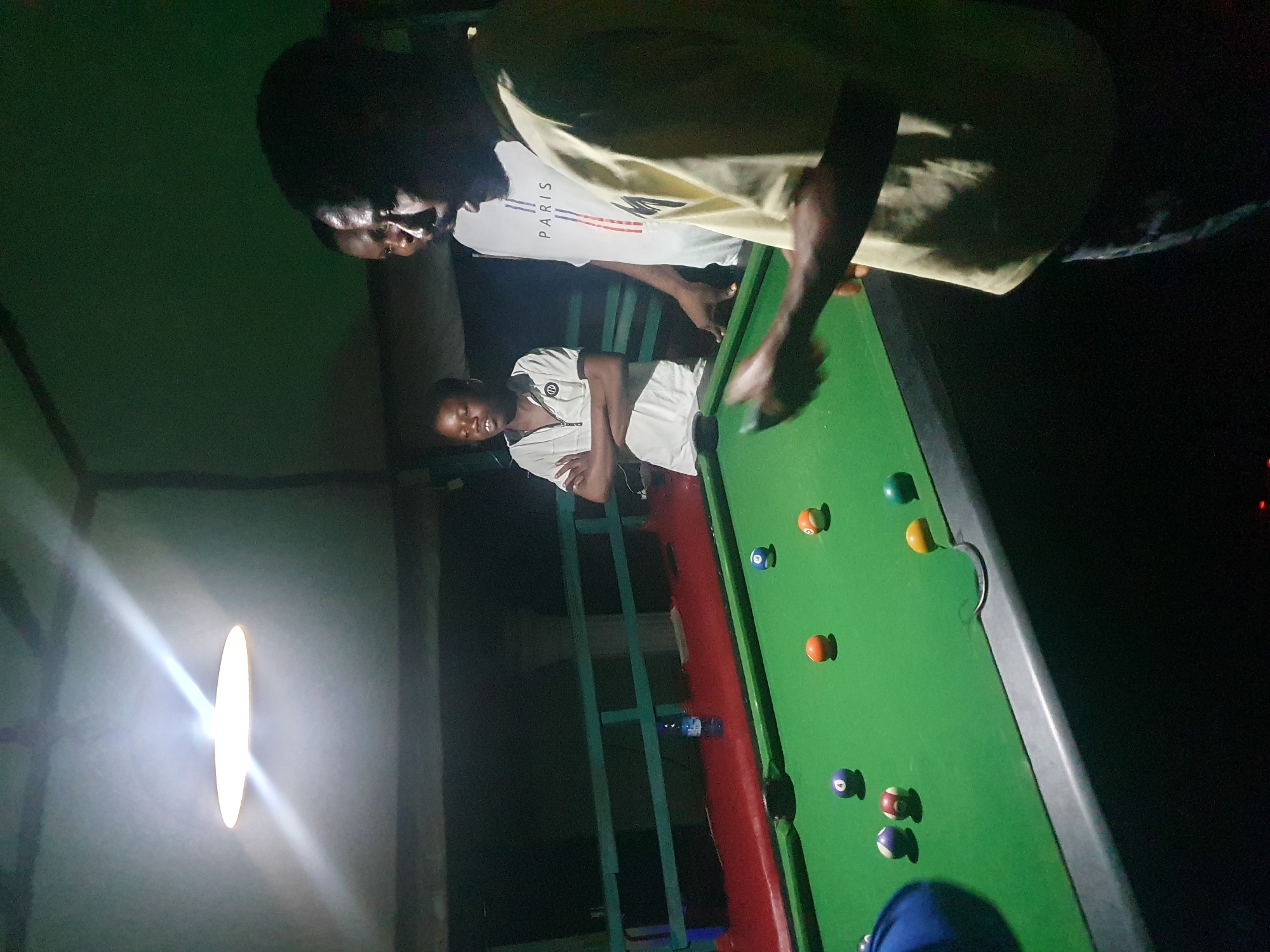 The image depicts an indoor billiards area, possibly a bar or recreational center, illuminated by a bright overhead light. Three young men are visible. In the foreground on the right, one man in a light-colored t-shirt is holding a cue stick, appearing to be in the process of playing. Another man, wearing a white t-shirt with "PARIS" printed on it, stands behind him and to the left. A third, younger individual stands further back on the left, with his arms crossed. A green billiards table occupies the lower portion of the frame, with several colored balls scattered on its surface. A blue water bottle rests on a ledge behind the table, which is bordered by a red edge and a green railing. The background is dark, suggesting it is nighttime or the area is dimly lit. The setting is likely in Lokoja, Nigeria, given the context provided.