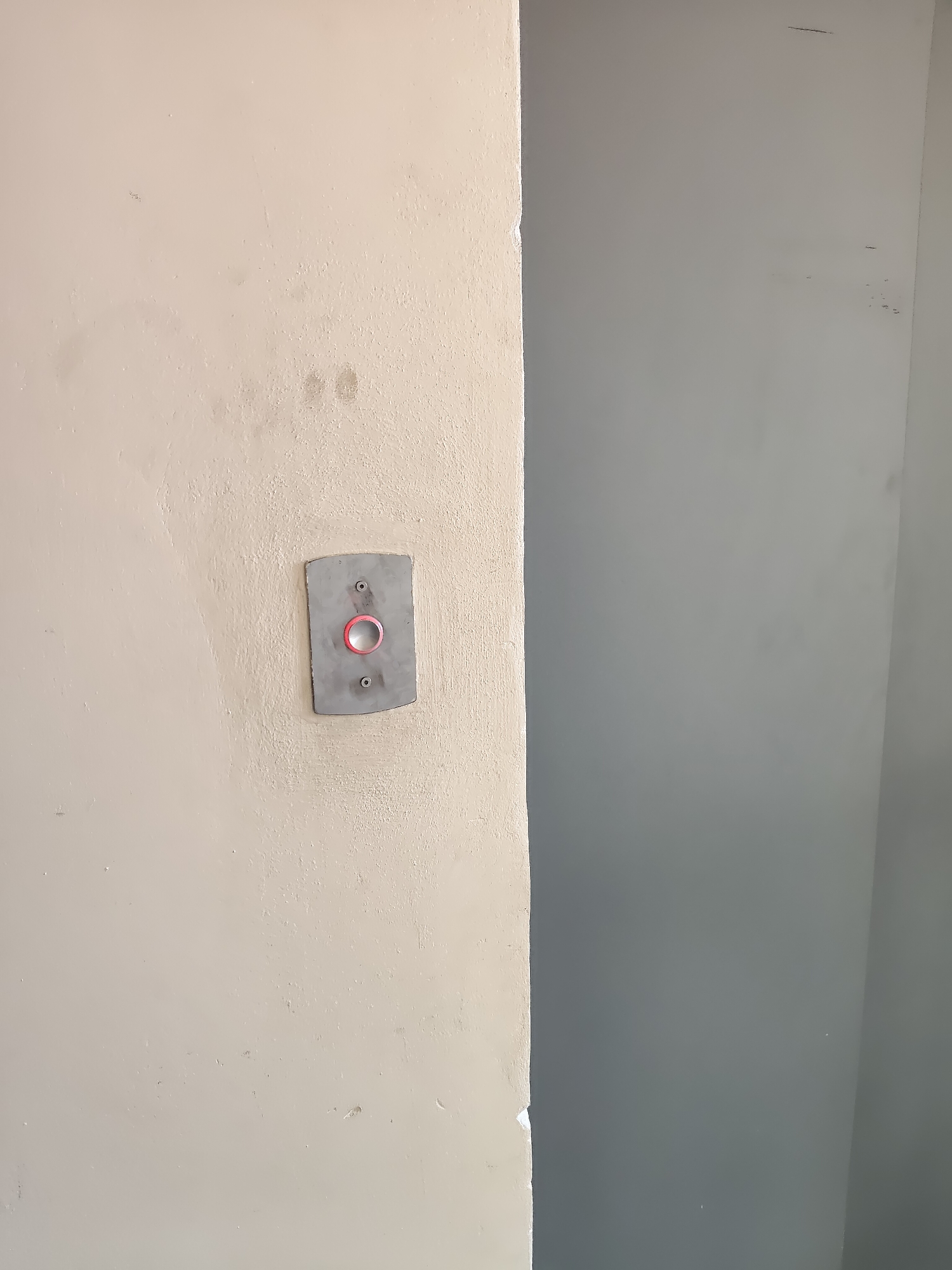 The image shows a close-up of a wall with an elevator call button. The wall on the left is a textured beige color, and the wall on the right is a smooth gray. The elevator call button is mounted on a gray metal plate that is attached to the beige wall with two screws. The button itself is a round, pink-colored ring surrounding a dark center. The scene appears to be indoors, likely in an apartment building or office. The image does not provide any specific information about its location in the city of Ungheni, Moldova.