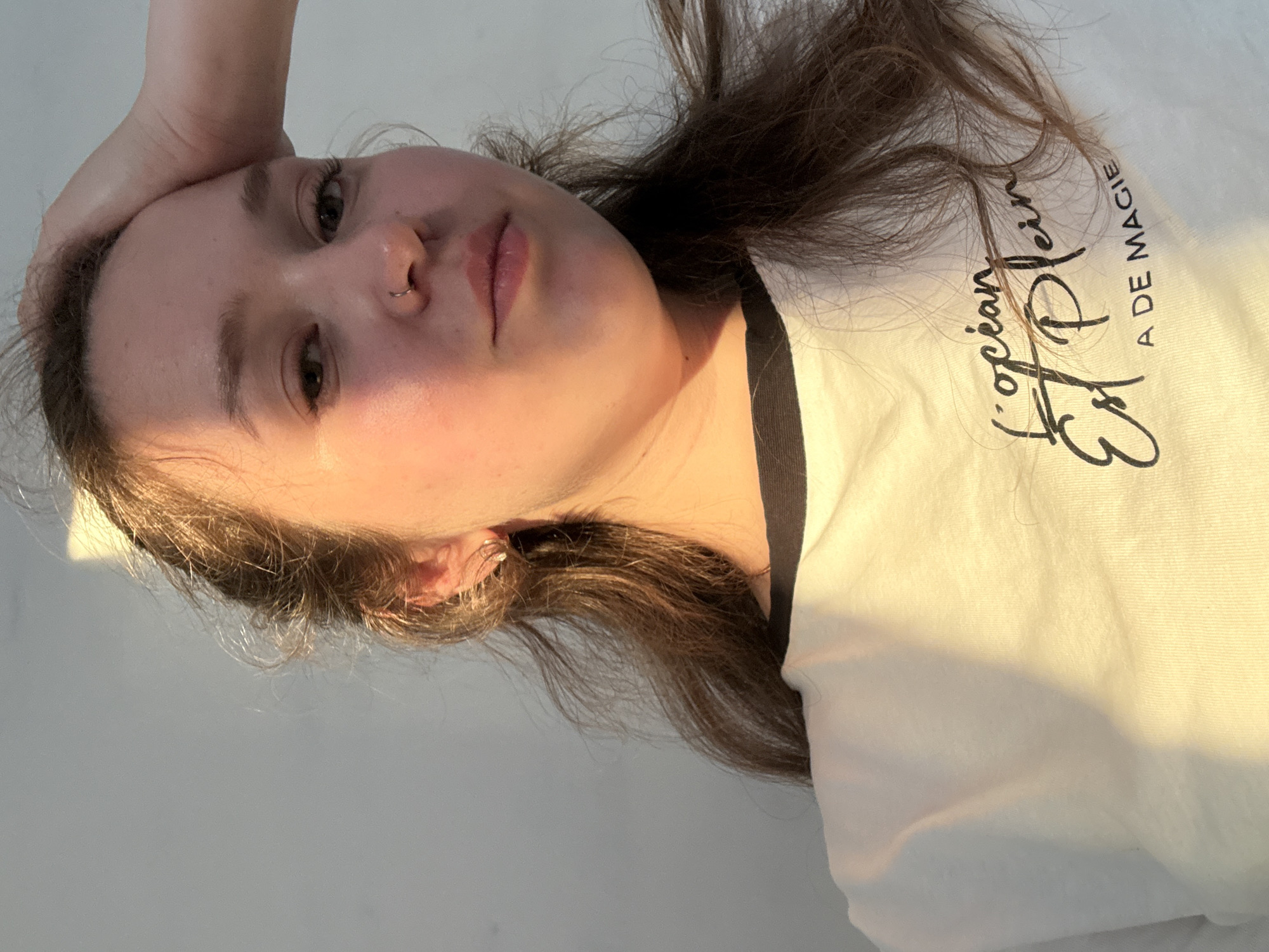 A young woman with long brown hair is lying on a white surface. Her left hand is on her forehead. She has a nose ring. She is wearing a white T-shirt with black trim and the words "L'océan Est Plein A DE MAGIE" written on it. The lighting is warm and appears to be natural. The background is a plain white surface. No location information is given.
