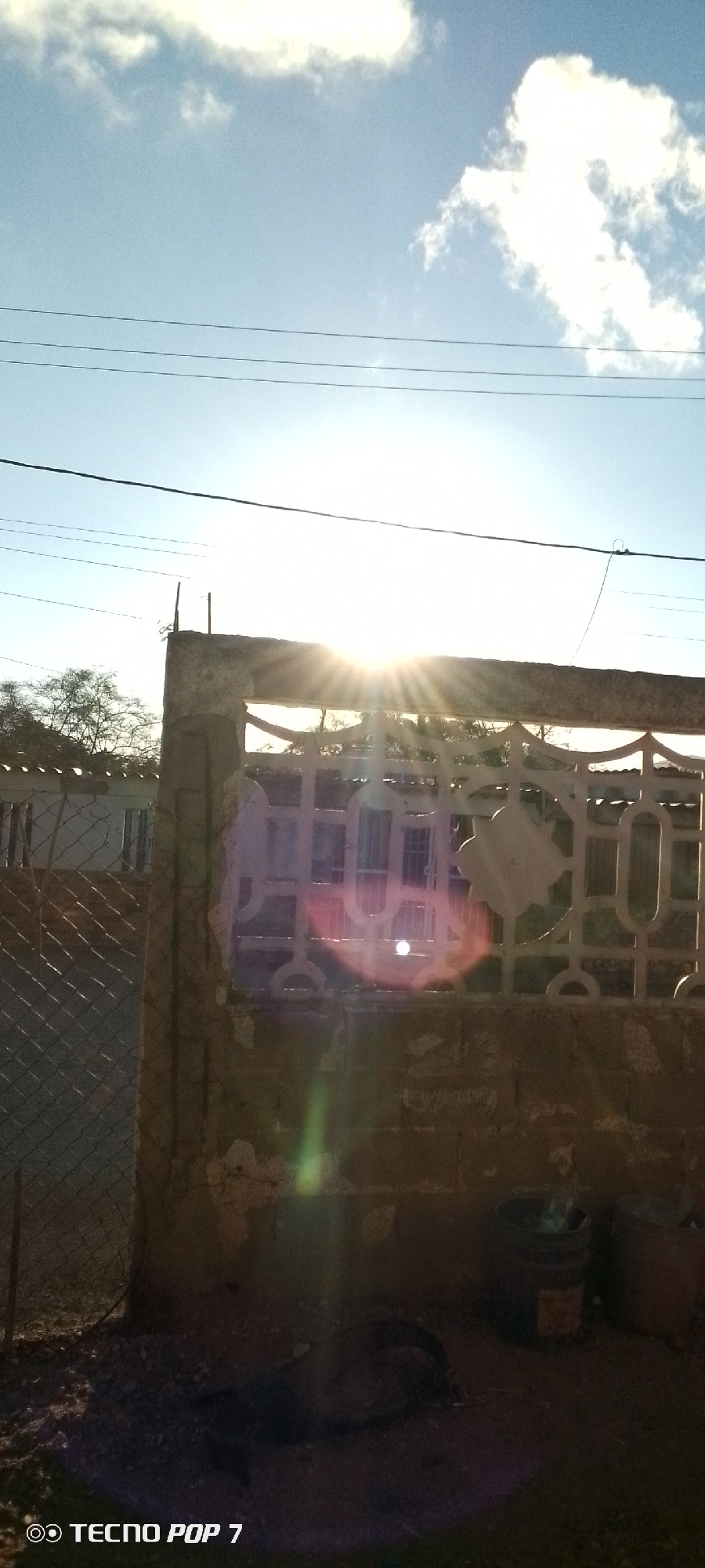 The image depicts an outdoor scene in Altagracia, Venezuela. The sun is high in the sky, partially obscured by clouds, and positioned directly behind a concrete and metal fence, causing lens flare. Power lines are visible against the sky. The fence is weathered and partly damaged. A chain-link fence and a building with white walls are visible in the background. On the ground near the fence are dark, indistinct objects, including what appear to be a tire and buckets. A "Tecno Pop 7" watermark is at the bottom left corner of the image.