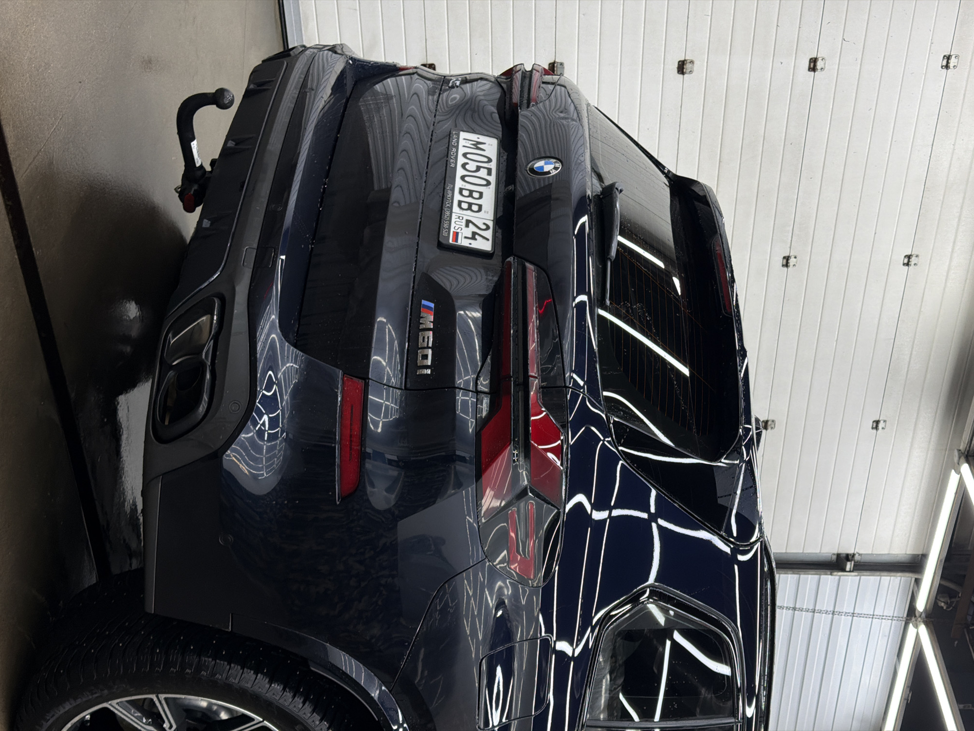 A close-up shot captures the rear of a dark blue BMW SUV in a car wash setting. The vehicle is positioned against a backdrop of a white, horizontally-paneled wall, reflecting the clean, polished surface of the car. The SUV's license plate, "M050 BB 24", is prominently displayed, with details identifying it as registered in Russia, specifically in the Irkutsk region, with a phone number listed below (3952) 550-520. A tow hitch is visible on the left side of the frame, and the tire suggests it is a large SUV. The car is angled, highlighting its taillights and rear window, which reflects the overhead lighting of the car wash. The reflections and the setting indicate a recent or ongoing cleaning process.