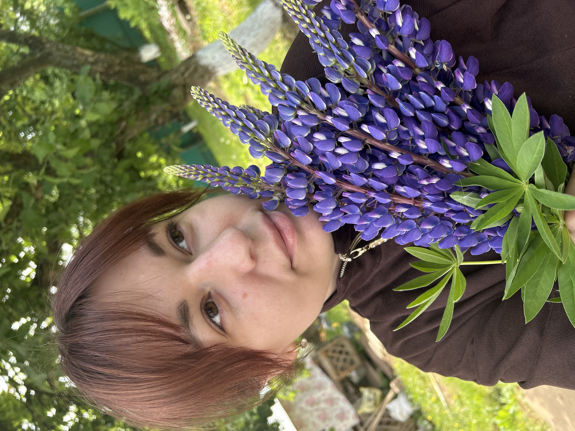 A rotated close-up shot features a woman holding a bouquet of purple lupine flowers. The woman has fair skin, brown eyes, and short reddish-brown hair. She is wearing a dark brown, possibly knitted, top and a necklace with a gold chain. The lupine flowers are densely packed together with some green leaves visible. The background is out of focus, showing green foliage and hints of a wooden bench or structure. It is not possible to determine the location as Фоки, Russia, with the given image content.