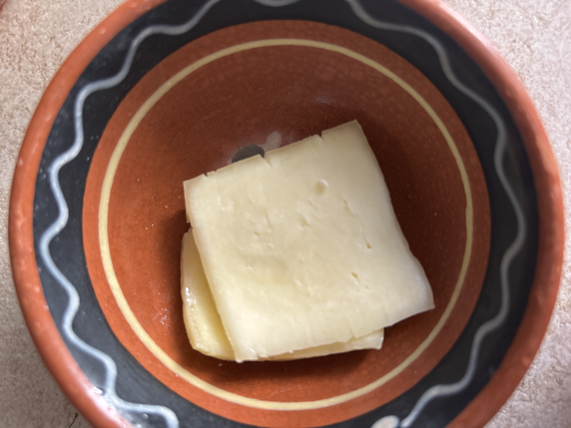 Two slices of light yellow cheese are stacked in an earthenware bowl. The bowl has a rustic, reddish-brown interior with concentric bands of yellow and black, the black band featuring a wavy white line. The bowl sits on a textured, light-colored surface. The scene is lit from above, casting a slight shadow. There is no indication of location within the city of Bila Tserkva, Ukraine.