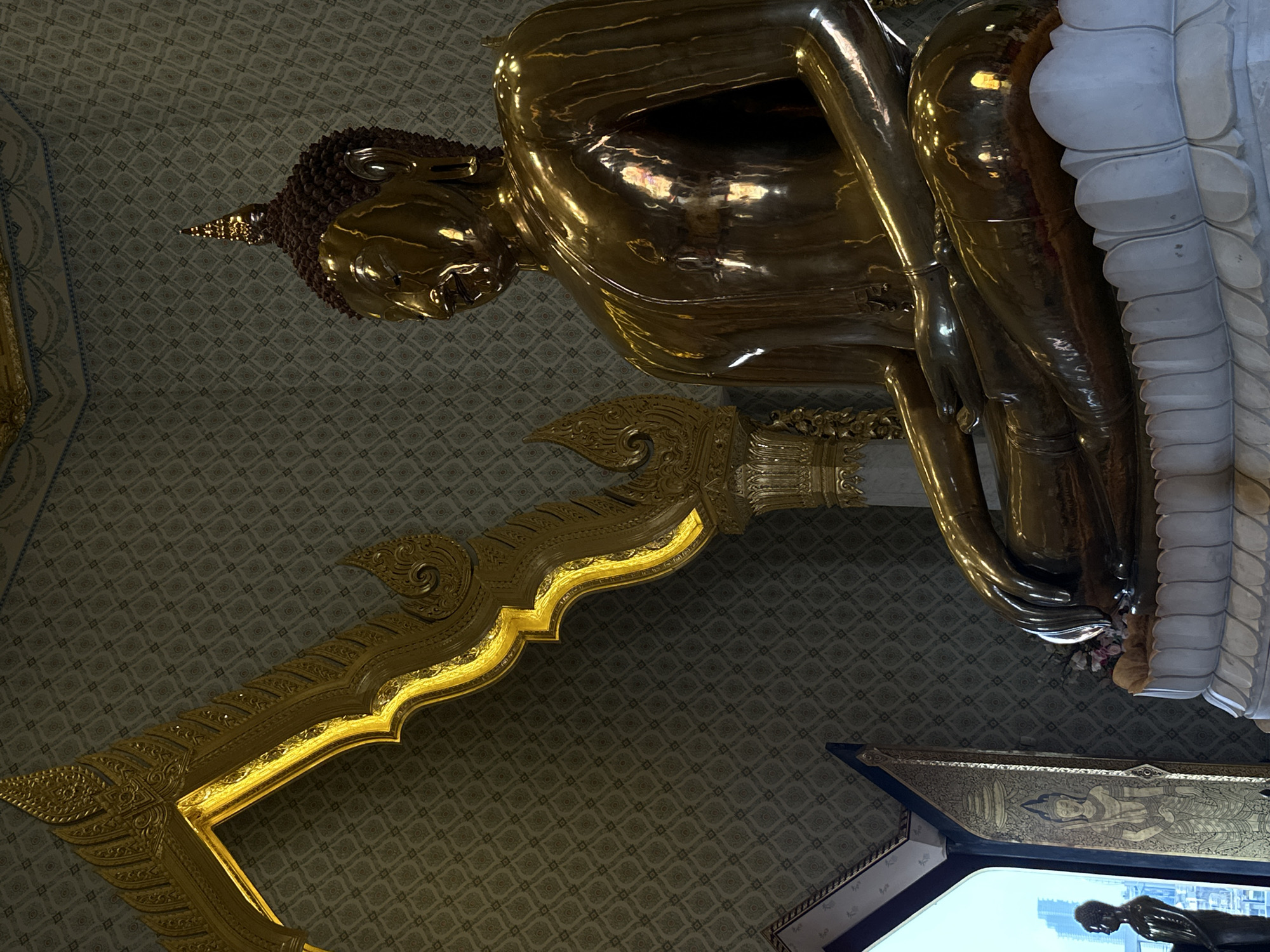 The image depicts a large golden Buddha statue, possibly at Wat Traimit in Bangkok, Thailand, based on the statue's appearance. The Buddha is seated on an ornate, golden platform. The backdrop is a patterned wall. Parts of the surrounding temple architecture are visible.