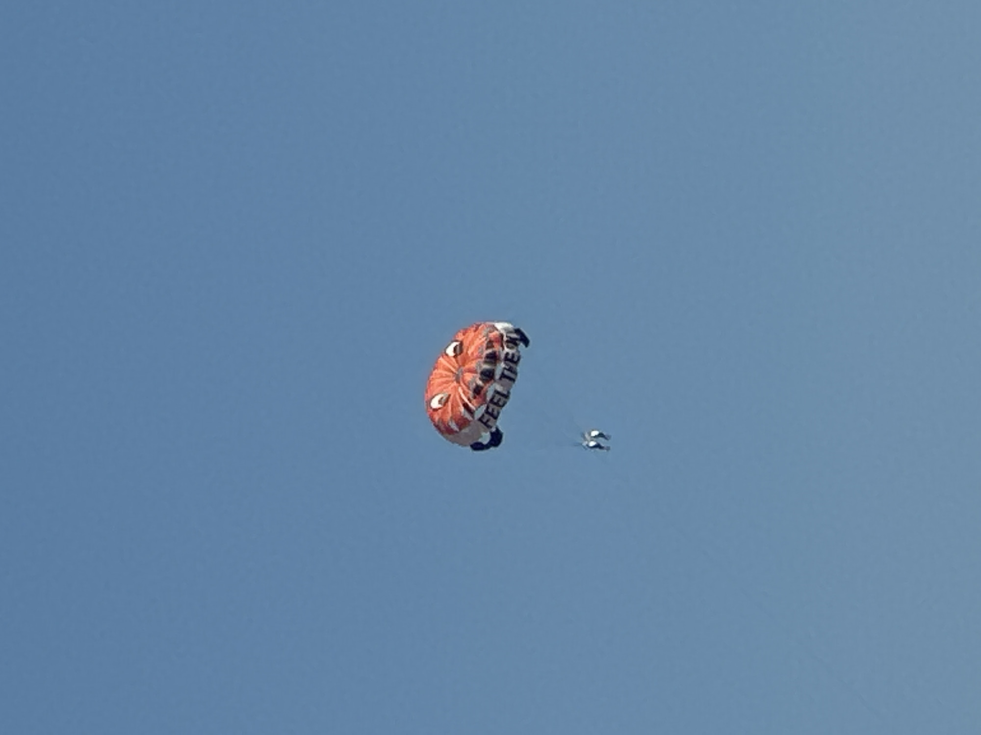 A parasail with a cartoon face design, colored red and white with black accents, is in mid-air against a clear blue sky. The text "FEEL THE" is visible on the underside of the parasail. Two figures are attached to the parasail by lines and are suspended beneath it. The scene takes place over the waters near Boreti, Montenegro.