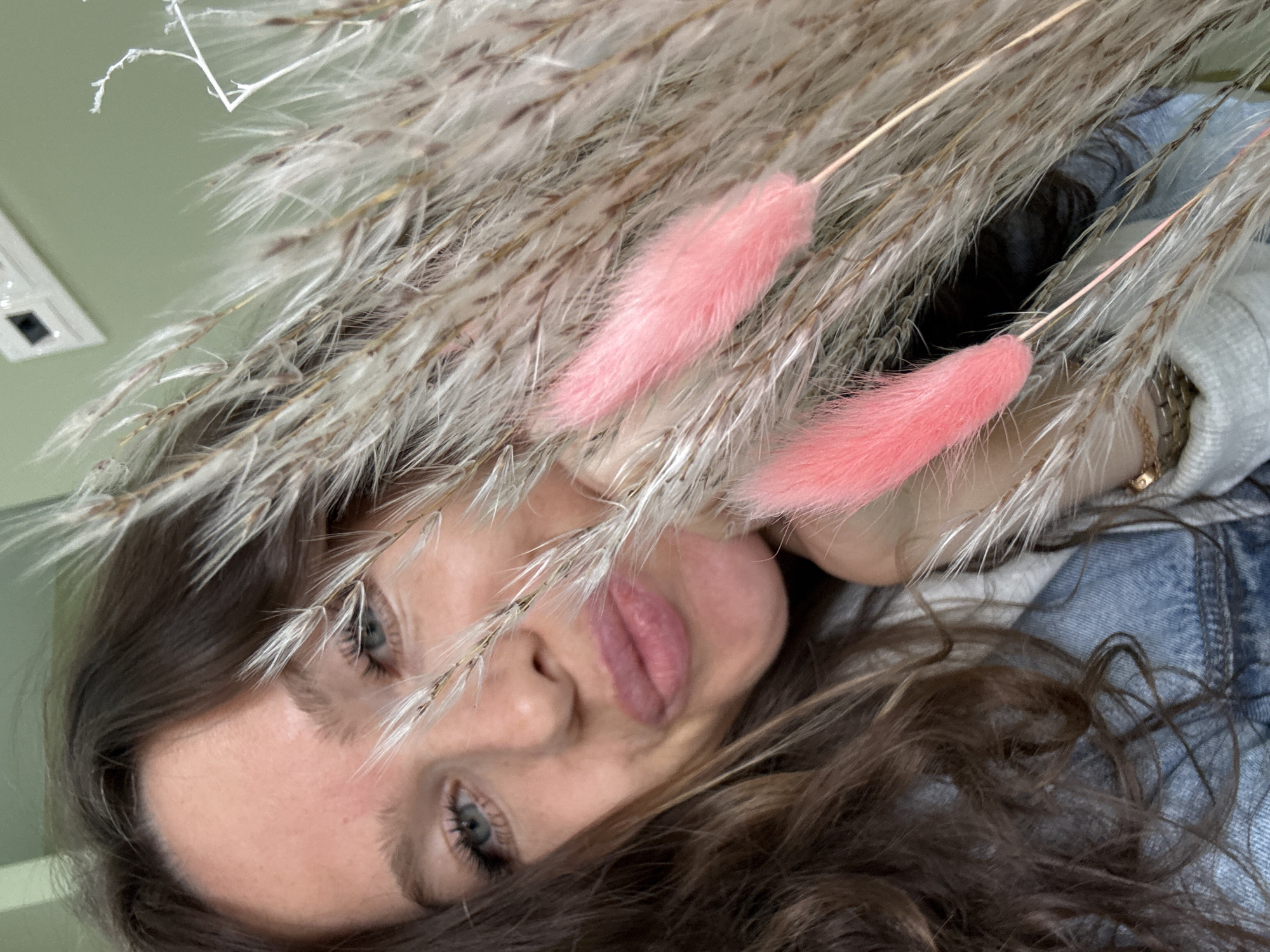 In Moscow, Russia, a young woman with long brown hair and blue eyes is captured in a close-up selfie. She is wearing a denim jacket over a grey hoodie. The woman is holding a bouquet of dried grasses and two fluffy pink bunny tails in front of her face, obscuring part of her features. Her lips are slightly parted, and she is looking directly at the camera. The background is a plain pale green wall with a light fixture visible at the top. The lighting is soft, suggesting an indoor setting.