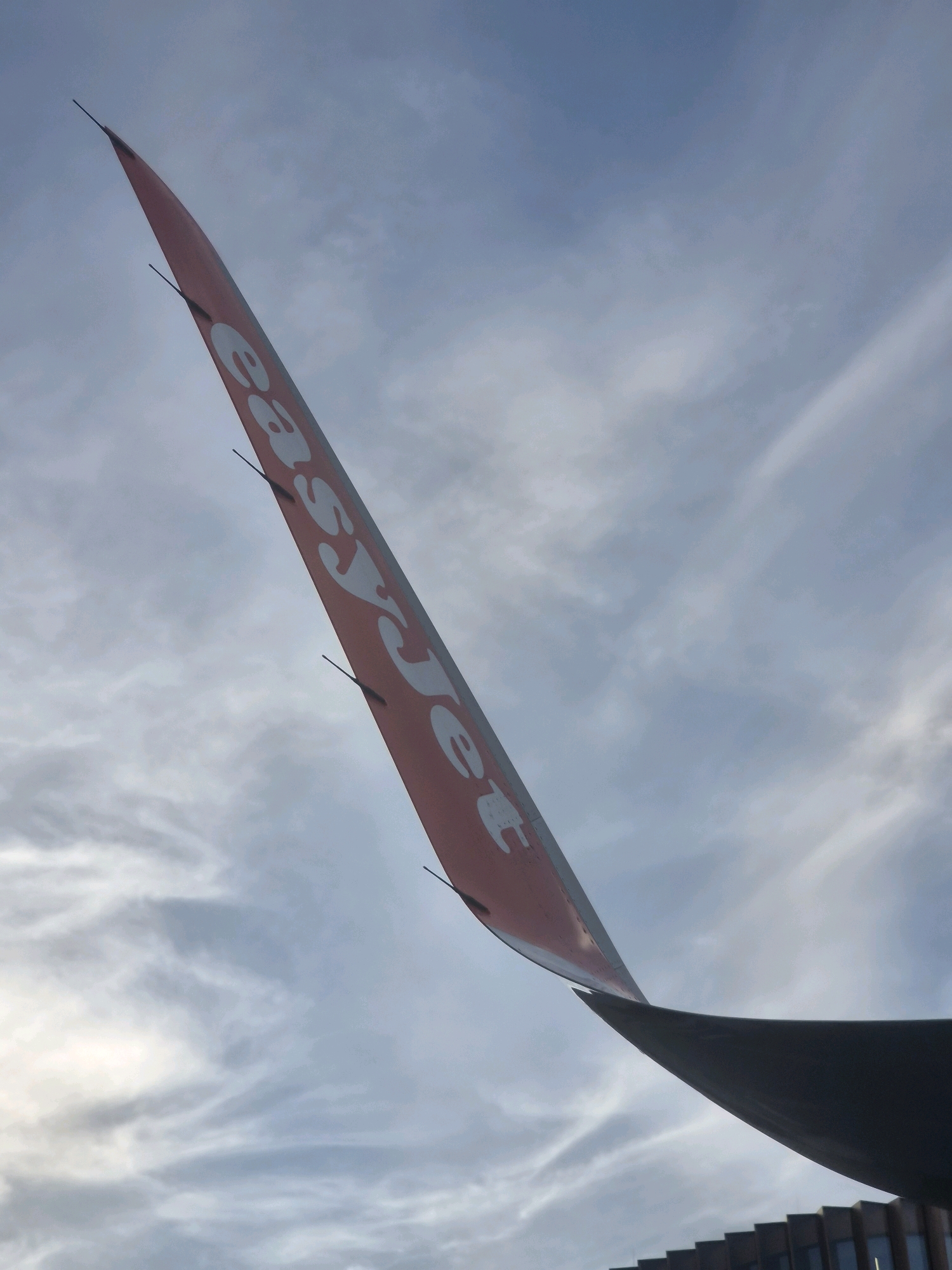 The image shows the wingtip of an airplane with the "easyJet" logo visible. The wingtip is angled upwards against a cloudy sky. The lower part of the image reveals the edge of another wing and a building with a dark, textured facade and horizontal window lines. The scene is outdoors, under a partially overcast sky, with the lighting suggesting it could be late afternoon or early morning. The specific location is the apron area of Luxembourg Airport (LUX) in Niederanven, Luxembourg. There are no people visible in the image.