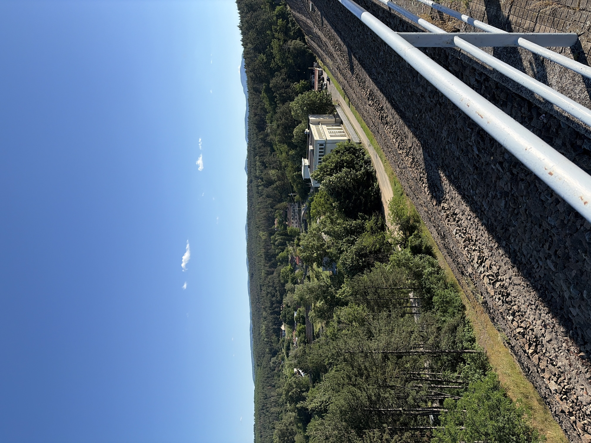 This is a landscape photograph taken on a clear, sunny day from a high vantage point, likely a dam or elevated walkway. In the foreground, a metal railing with white posts and horizontal bars runs diagonally from the bottom right towards the center of the frame. Below the railing, the ground is covered with a dense layer of dark, rough-textured stones, forming a steep slope. A diagonal shadow cuts across this rocky expanse, cast by the railing and the viewer's position.
Further down the slope, a paved road runs parallel to the railing. Alongside the road, a modern, rectangular building with numerous windows and a flat roof stands out. This building appears to be white or a very light color. Beyond the building, the landscape transitions into a dense forest of green trees that extends to the horizon. Within the forest, glimpses of houses and structures suggest a rural or semi-rural settlement. In the far distance, a range of hills or mountains forms the backdrop under a vast, clear blue sky with a few small, scattered white clouds.
The image captures the Koprinka dam area in Bulgaria, showcasing the dam's structure, the surrounding natural environment, and a glimpse of the adjacent infrastructure. There are no people visible in the image. The scene is peaceful and serene, with the sunlight highlighting the textures of the rocks and the vibrant green of the foliage.