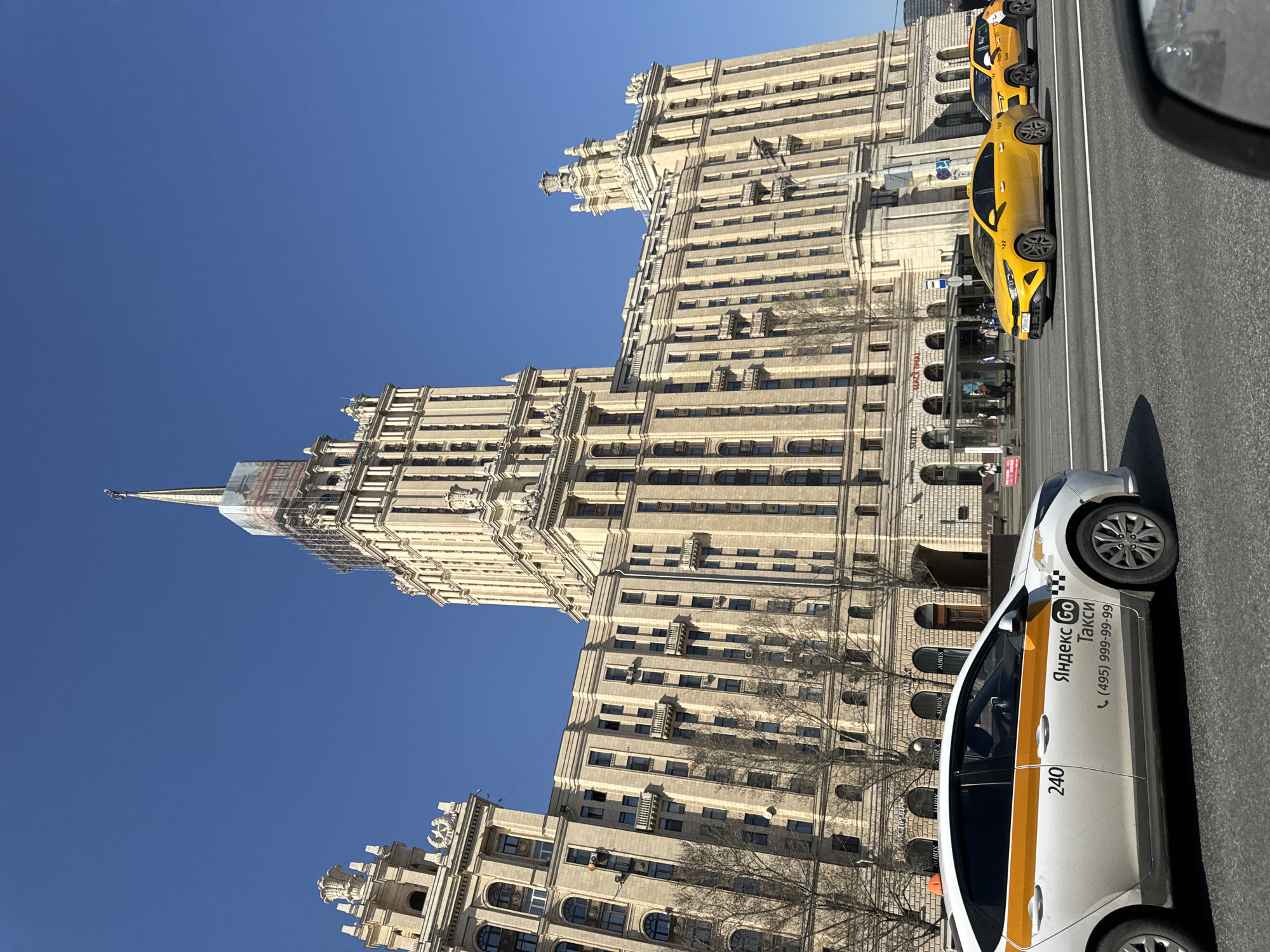 A tall building, likely one of the Seven Sisters skyscrapers in Moscow, Russia, dominates the scene under a clear blue sky. Scaffolding partially covers the top of the building's spire. A white Yandex Taxi vehicle with a yellow stripe is in the foreground, parked on the right side of the frame. Another yellow taxi is visible further down the street on the right. The building is light-colored, with many windows and architectural details. Bare trees are visible in front of the building. The scene appears to be taken from inside a vehicle, with a portion of the interior visible in the upper right corner.